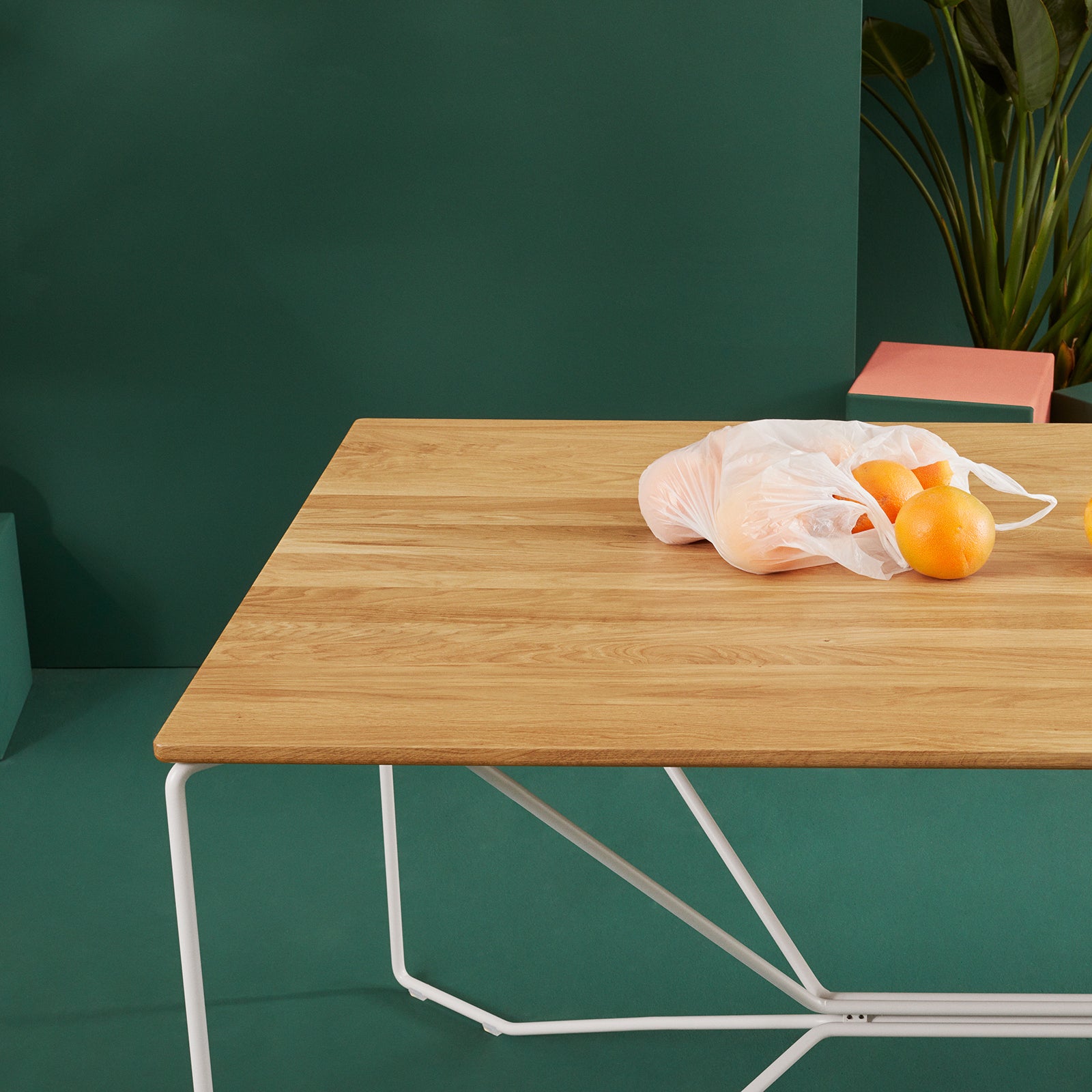 The SAME SAME Oak Table by Victor Foxtrot, featuring a powder-coated steel frame, is displayed against a green backdrop. A white net bag with oranges and a few scattered ones are on the table, while a small potted plant peeks from the background.