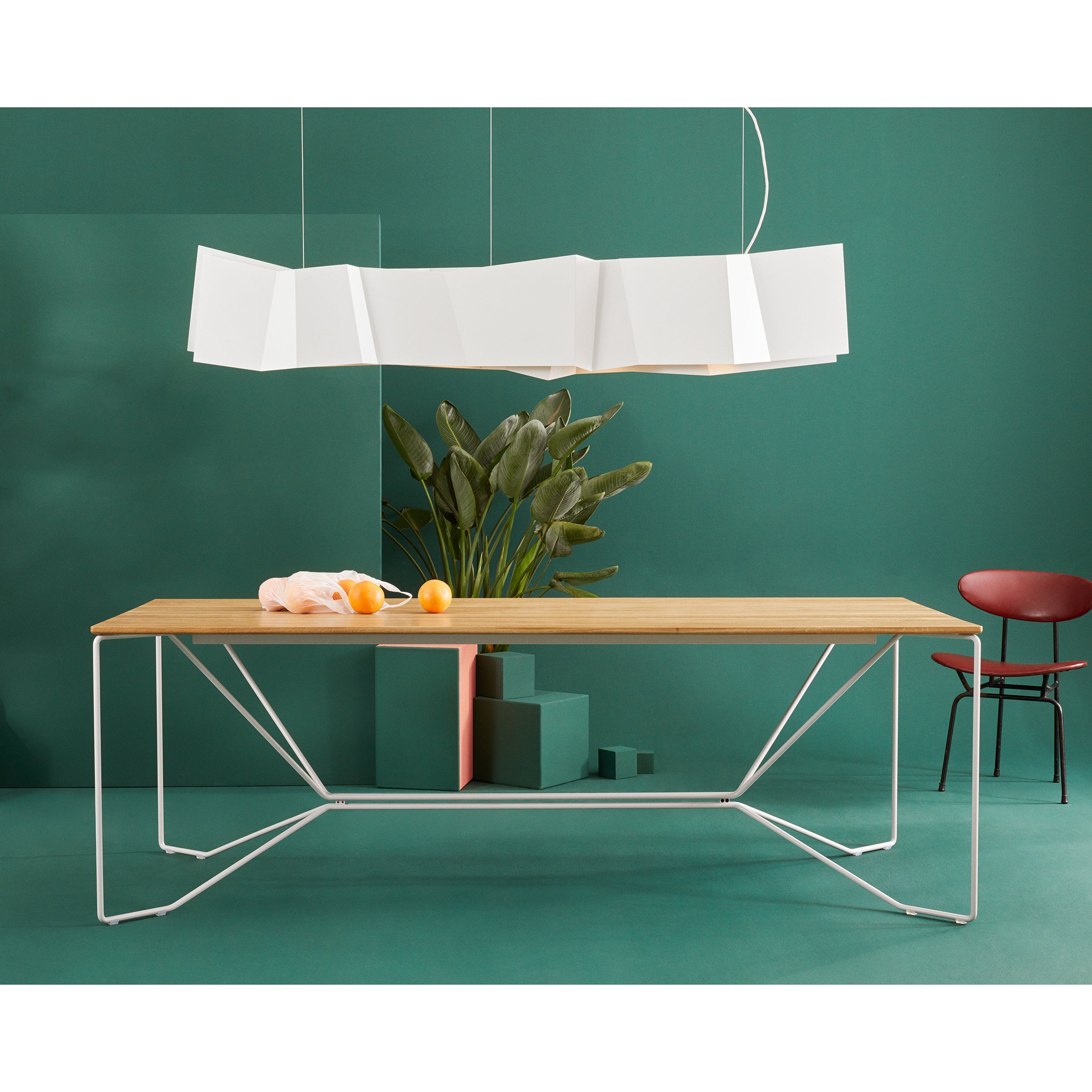 A modern dining area showcases the SAME SAME Oak Table by Victor Foxtrot, supported by a minimalist powder-coated steel frame. Its illuminated by a geometric white pendant light, with a red chair and green backdrop, accented with oranges on a pink cloth and potted plants.