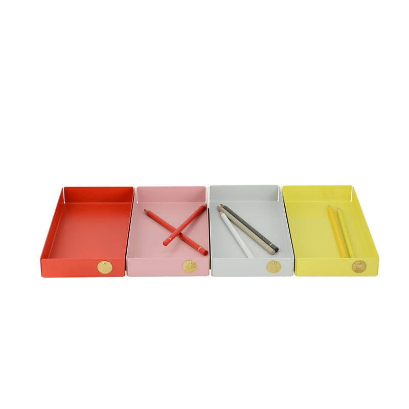 Four POI Tray M trays from Victor Foxtrot, designed for design lovers, are side by side in powder-coated steel. From left to right: an empty red tray, a pink tray with crossed red pencils, a light grey tray with two colored pencils, and a yellow tray with two yellow pencils. Each features a circular label.