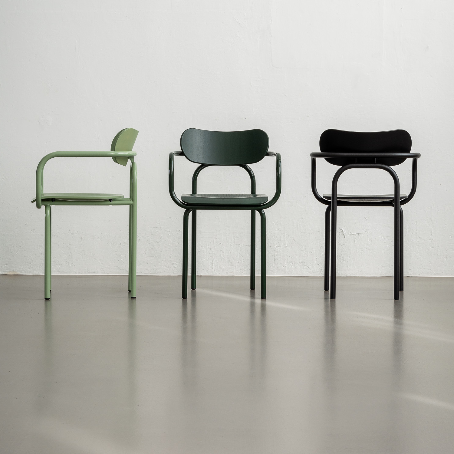 Three Victor Foxtrot Double U Armchairs stand on a smooth light gray floor. The left chair has pressed beech and light green armrests, the middle sports dark green with no armrests, and the right is black with tubular steel accents—all against a plain white wall.
