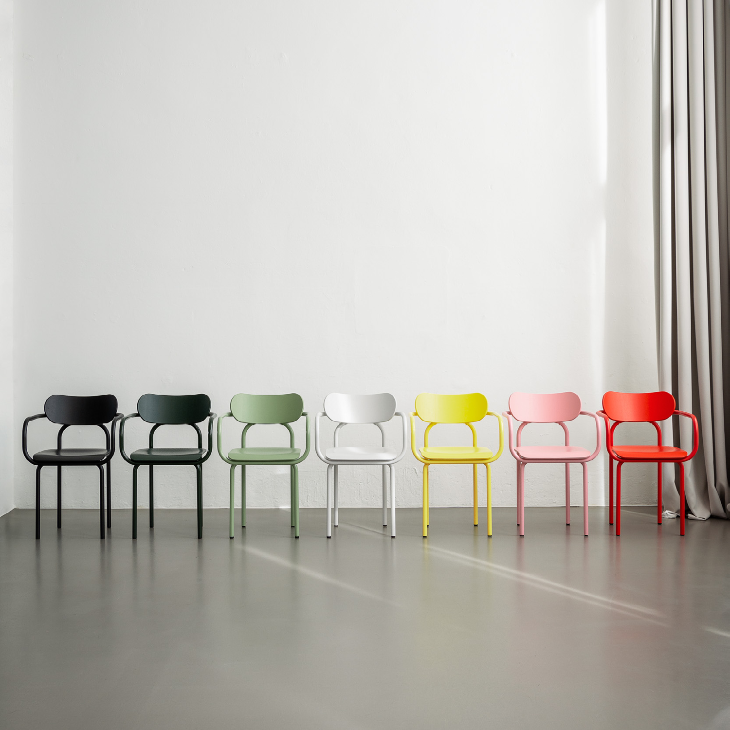 Seven iconic and colorful Double U Armchairs by Victor Foxtrot—black, dark green, green, white, yellow, pink, and red—stand against a white wall. They feature a blend of tubular steel and pressed beech. A gray curtain partially drapes the right side of the wall as it meets the gray floor.