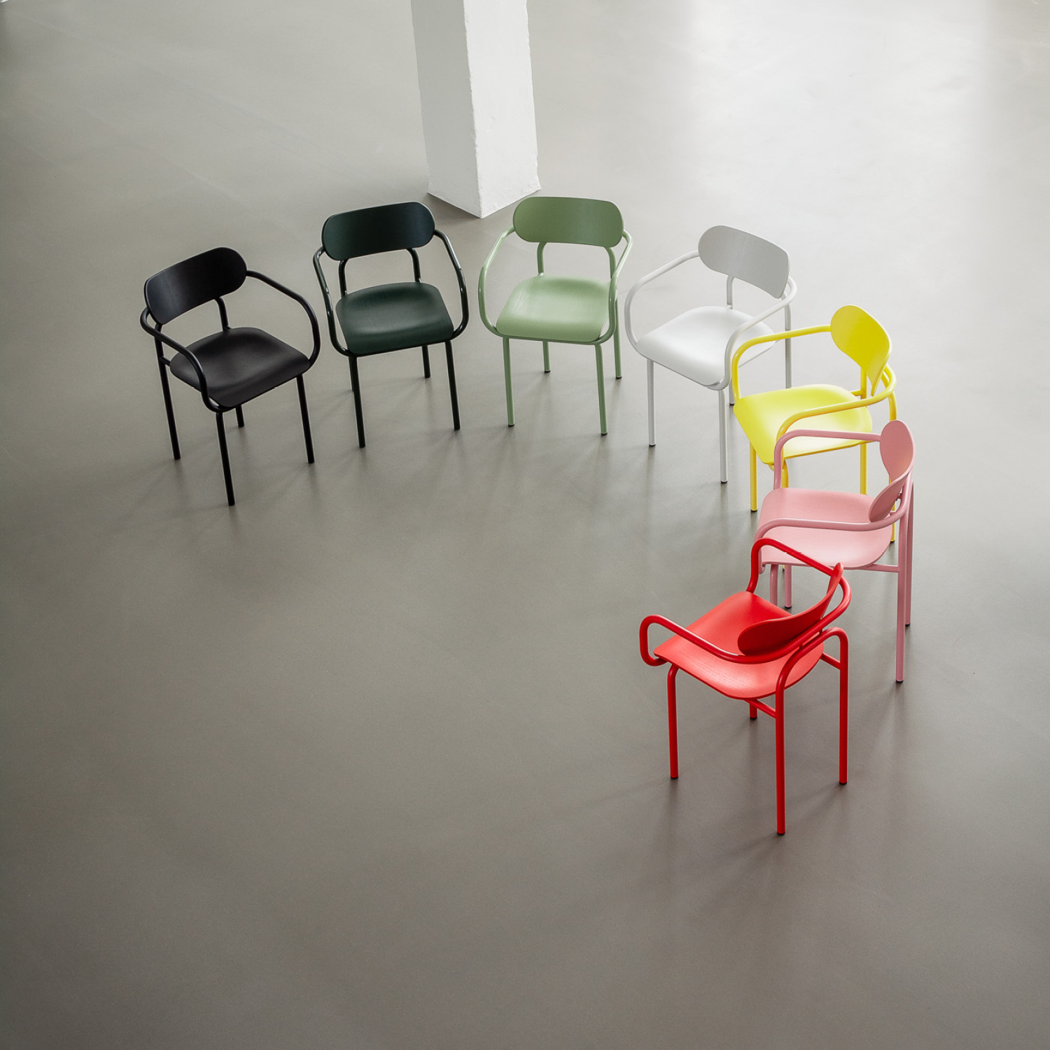 The Double U Armchair by Victor Foxtrot features seven tubular steel chairs in black, dark green, light green, white, yellow, pink, and red. They form a circle on a light gray floor within an otherwise empty room.