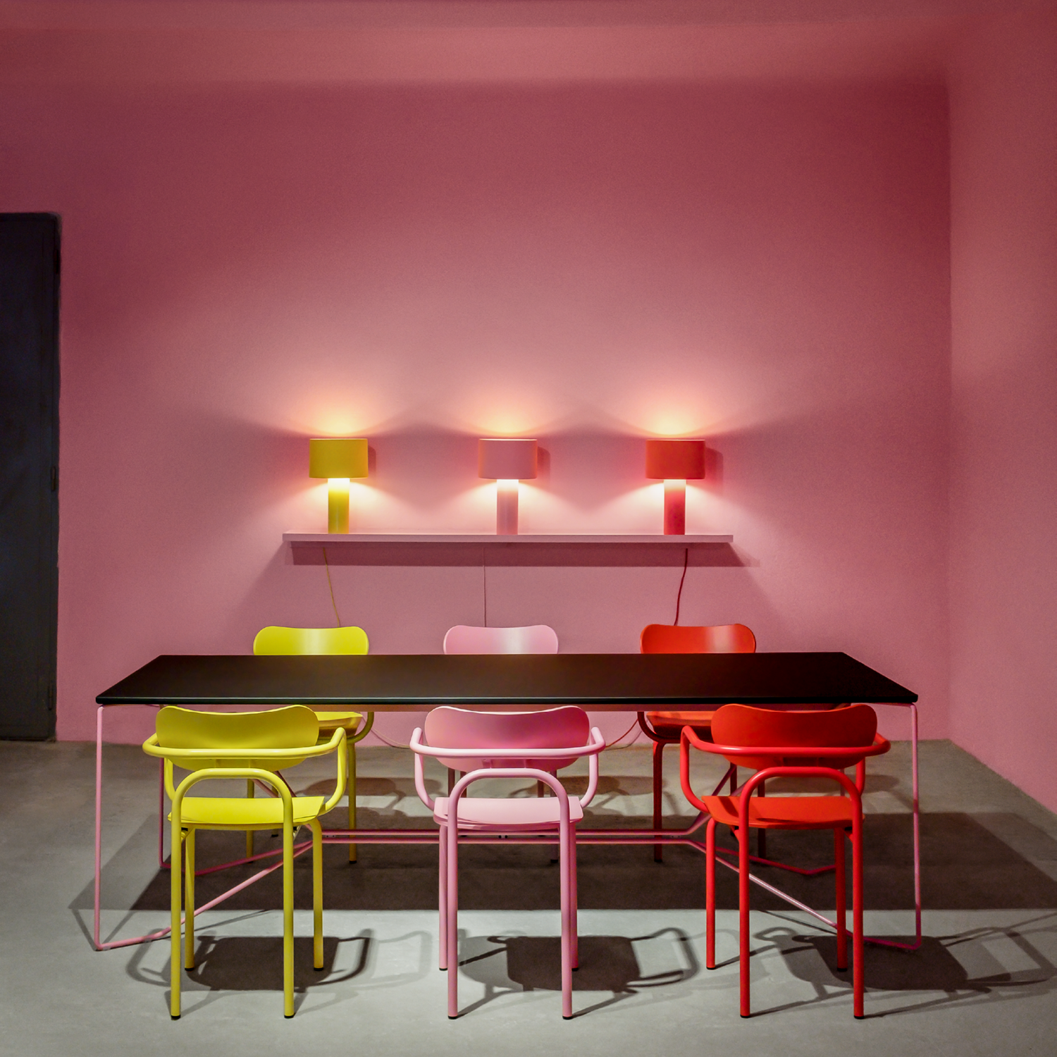 A modern dining room features a black table with vibrant Double U Armchairs from Victor Foxtrot in yellow, pink, red, and orange. The tubular steel frames complement pink walls with three red wall-mounted lamps for warm light, highlighting the bold and iconic aesthetic.