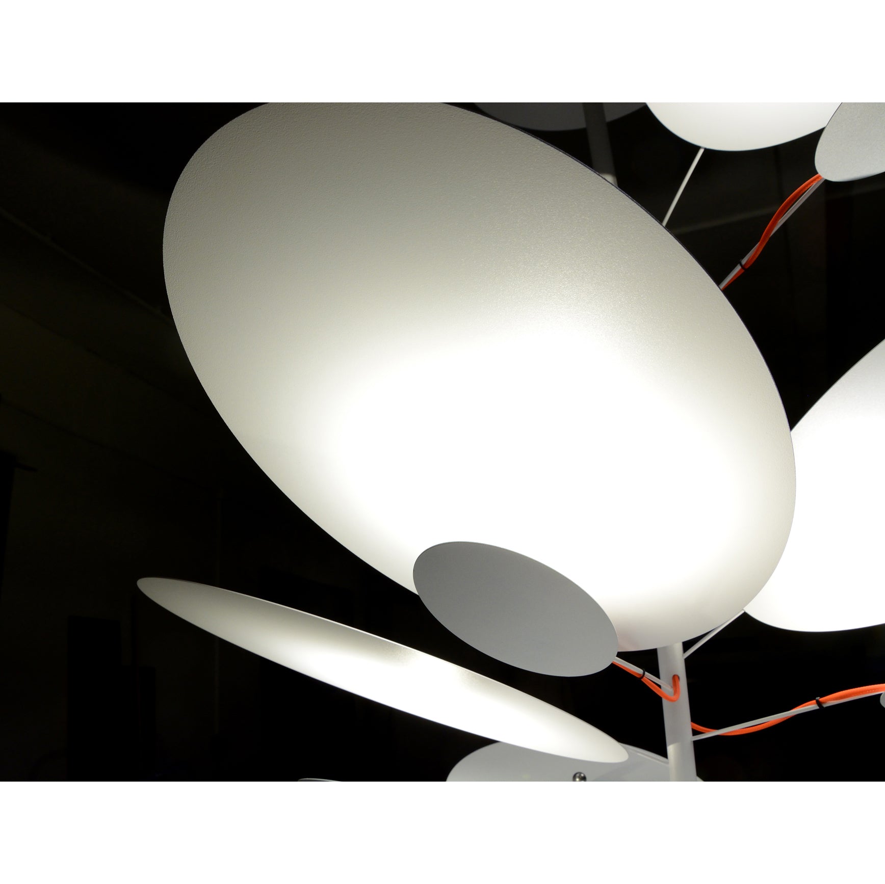 Close-up of the LEAF IT Pendant Lamp by Victor Foxtrot, showcasing modern pendant lights with overlapping round white discs that emit a soft glow in a dark room. Suspended on slim vertical rods, these lights embody an organic and minimalist design with an abstract appearance.