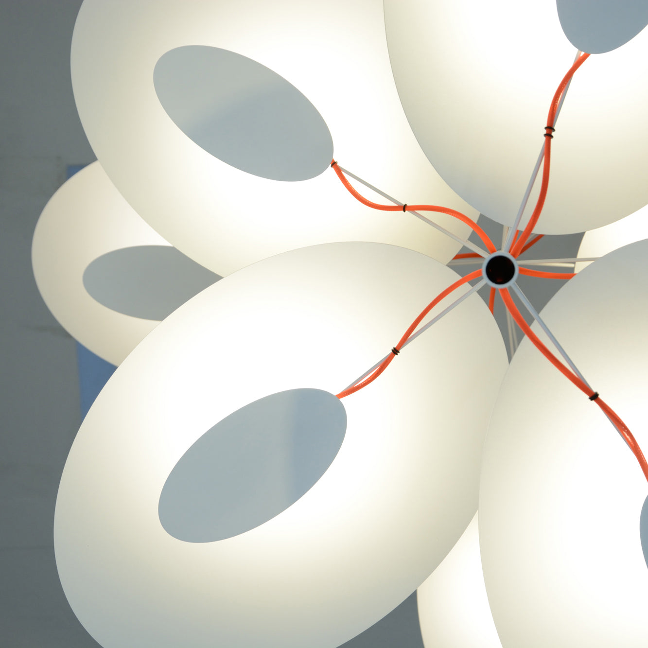 Close-up of the LEAF IT Pendant Lamp by Victor Foxtrot: white, oval lampshades arranged in a circle with red wires connecting to a central black hub, resembling reflective leaves. The softly lit background highlights its floral-like elegance.