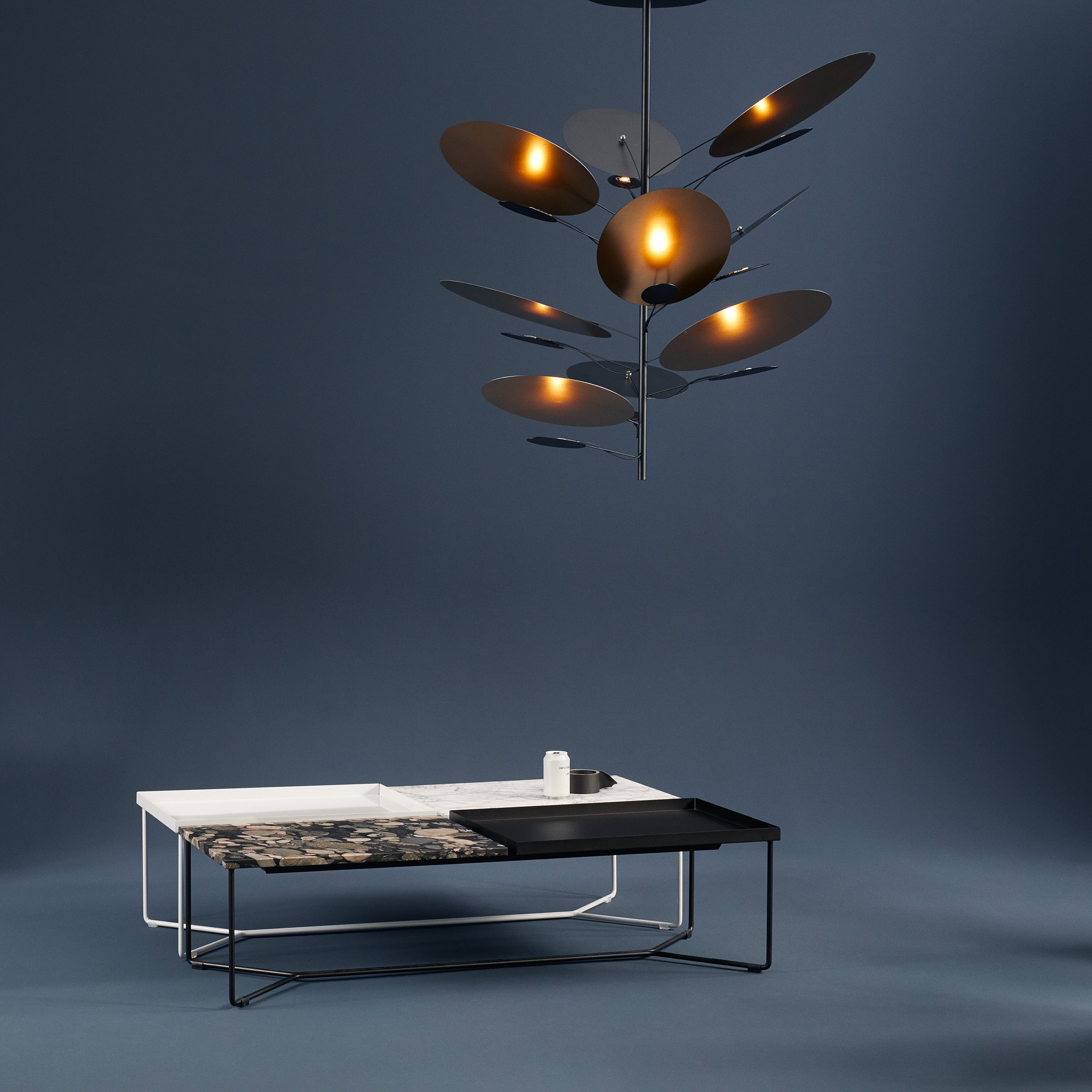 A sleek coffee table features two sections: one white with a candle and another dark with a pattern. Above, the Victor Foxtrot LEAF IT Pendant Lamp showcases organic, reflective leaves radiating warmth against a dark blue backdrop.