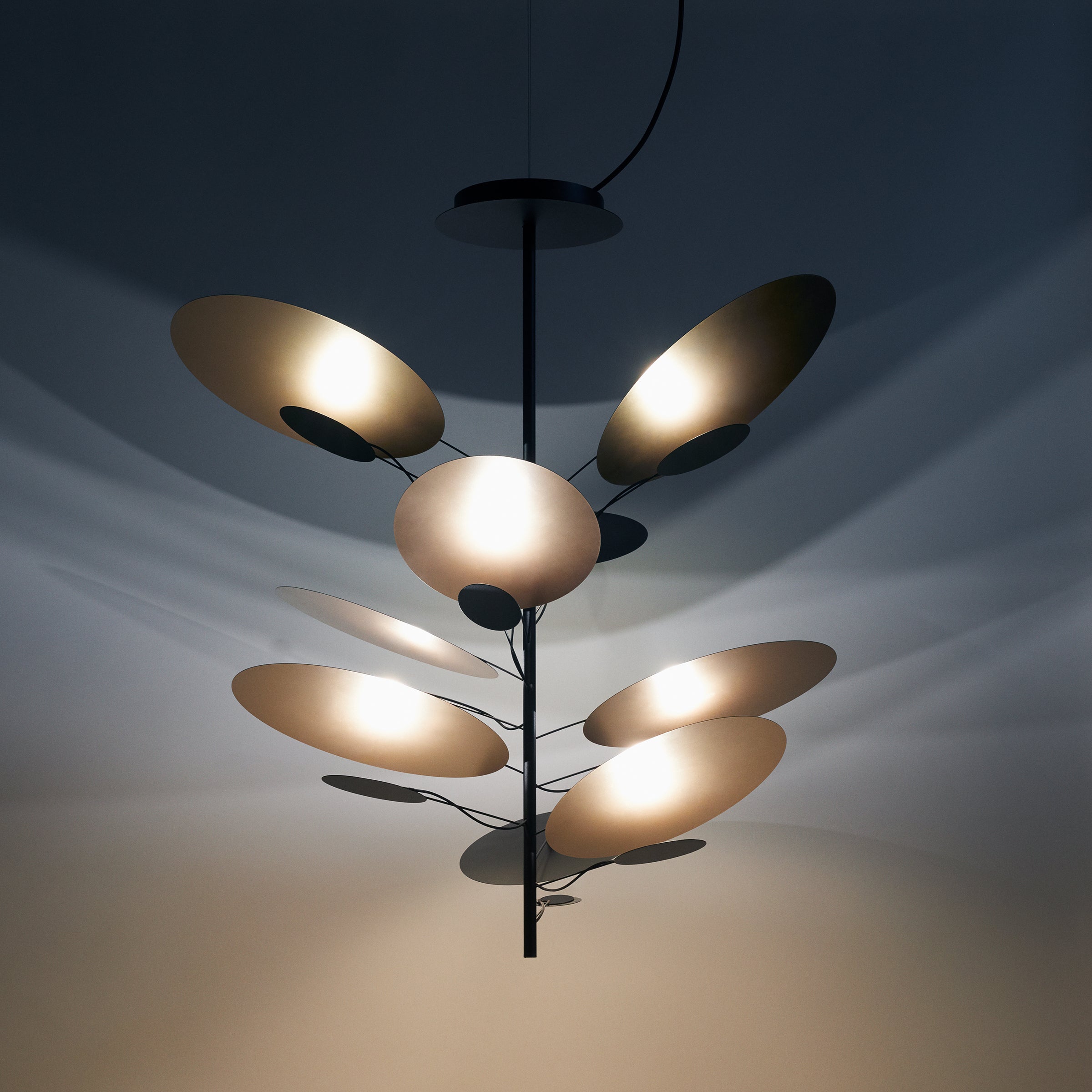 Victor Foxtrots LEAF IT Pendant Lamp features a modern chandelier design with a black central rod and multiple disc-shaped lights resembling reflective leaves, casting artistic shadows on the ceiling. The lamp’s organic design is perfectly complemented by plain walls.
