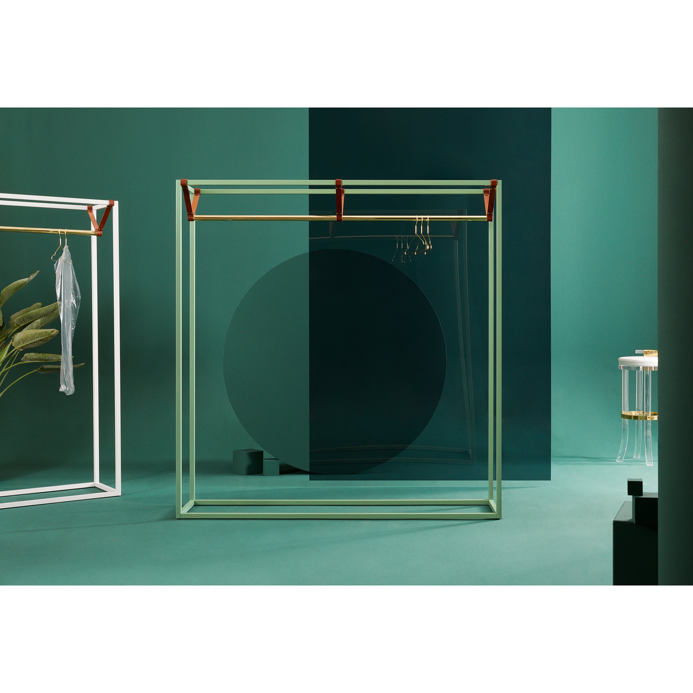 The minimalist space, featuring geometric frames and green walls, showcases a hanging garment from the Victor Foxtrot LESS MESS Coat Stand. On the right is a small table with glass decorations crafted from powder-coated steel. A large semi-transparent circle elegantly complements the green back wall.