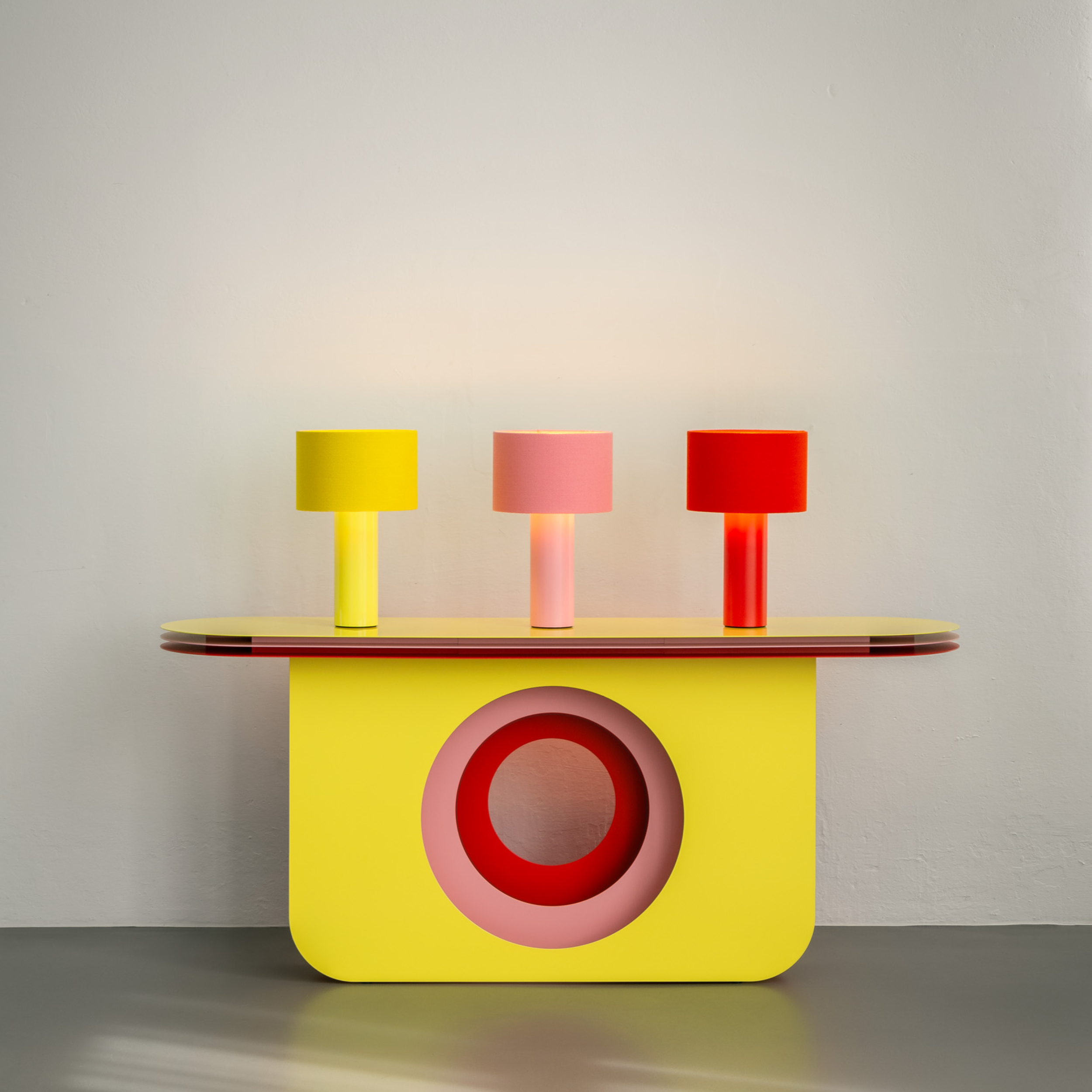 A modern My Ami Sideboard by Victor Foxtrot with a yellow circular powder-coated steel base and a smooth, rounded top. Three lamps in yellow, pink, and red sit on it, enhancing its depth against the plain light wall.