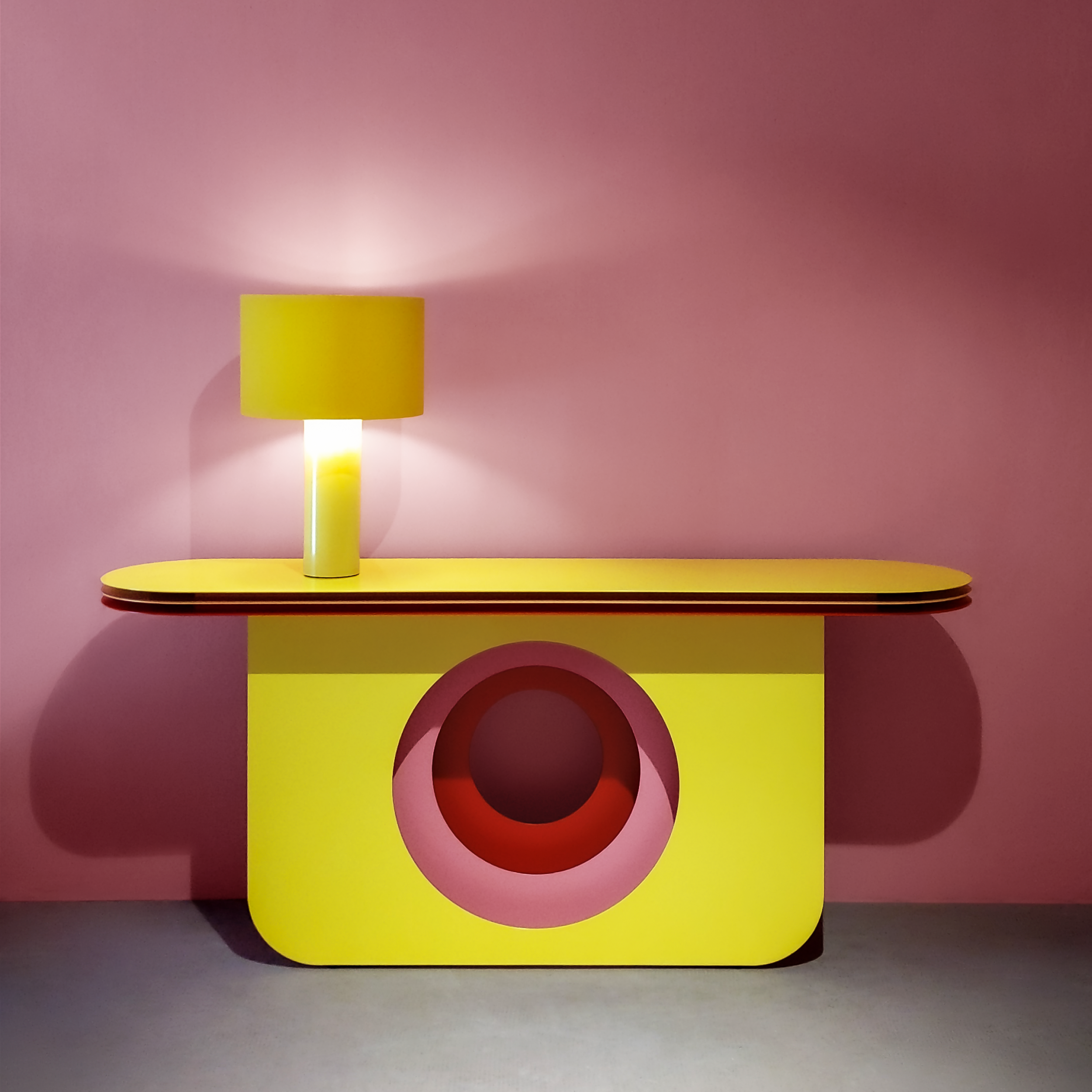 The My Ami Sideboard by Victor Foxtrot, a bright yellow console, stands against a pink wall. Its crafted from powder-coated steel and features a circular cutout. A matching lamp with a cylindrical shade complements it, casting soft shadows on the gray floor.