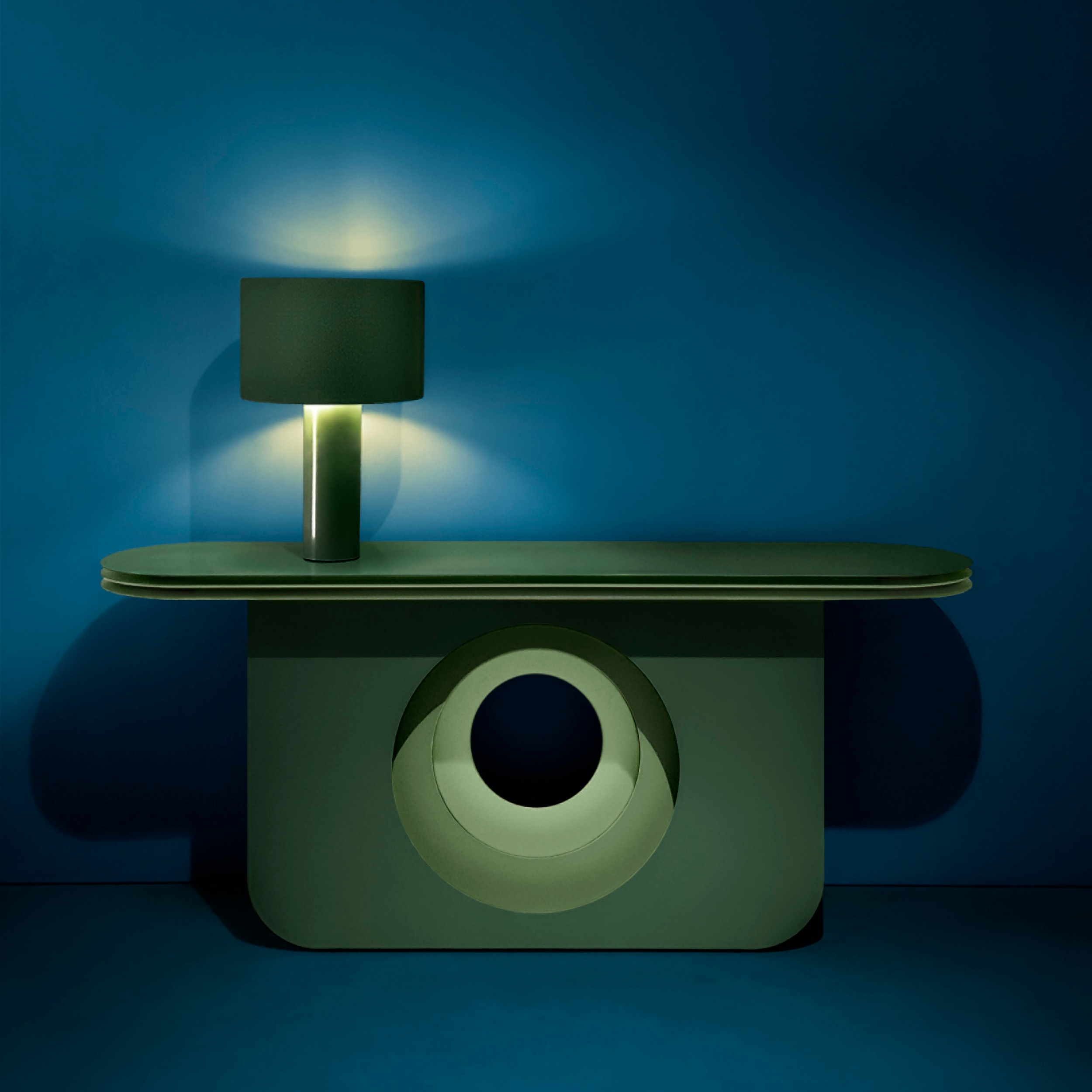 The My Ami Sideboard by Victor Foxtrot showcases a geometric design and an open circular cutout against a dark blue backdrop. Made of powder-coated steel, it pairs elegantly with a cylindrical lamp, its dark shade casting warm visual depth on the wall.