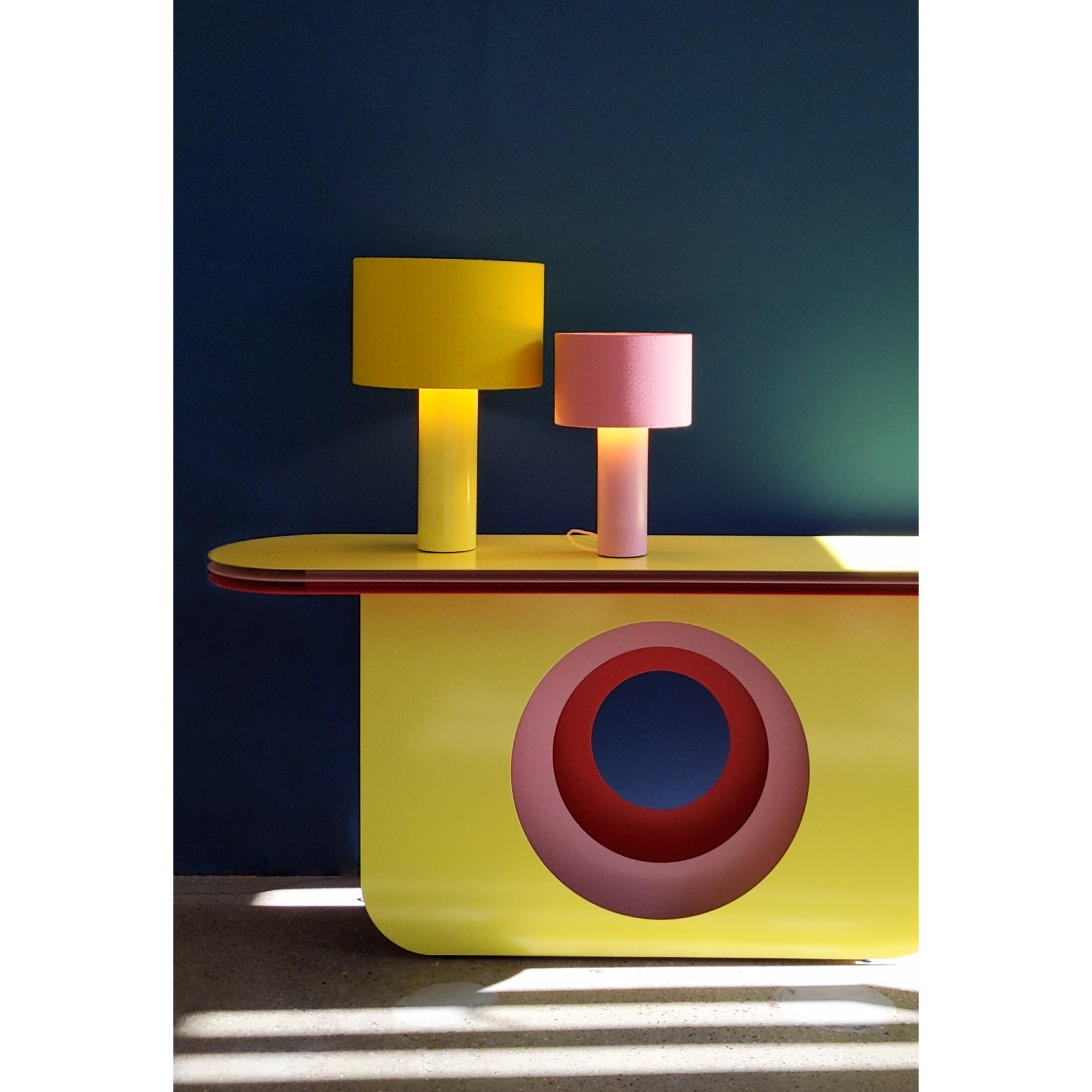 The My Ami Sideboard by Victor Foxtrot, crafted from powder-coated steel, features a large yellow and a smaller pink lamp. The yellow table boasted a circular pink and red design. Sunlight against the deep blue wall added visual depth to the scene.