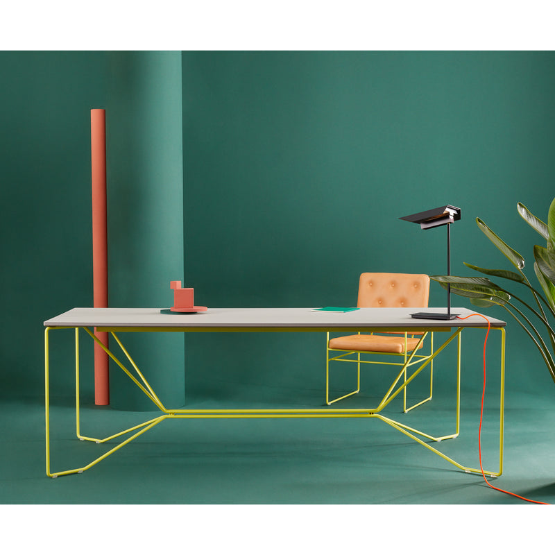 A modern desk setup features the SAME SAME Linoleum Table by Victor Foxtrot with a white top and yellow, powder-coated steel wireframe legs against a green background. An orange chair sits behind it with a notebook, lamp, red mug, plant, and tall pink cylinder completing the scene.