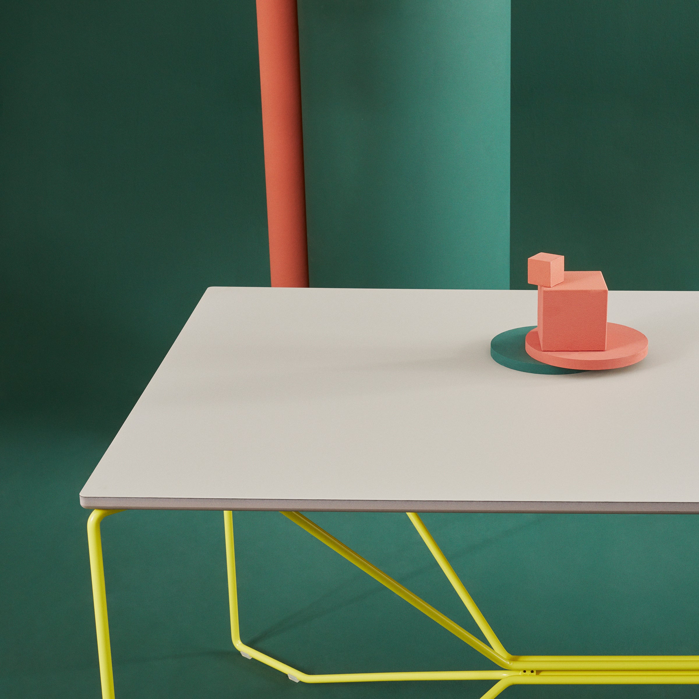 The image features the SAME SAME Linoleum Table by Victor Foxtrot, with a sleek white top and powder-coated yellow legs, set against a green and orange backdrop. On the table is a small abstract pink geometric sculpture with a matching pink base.
