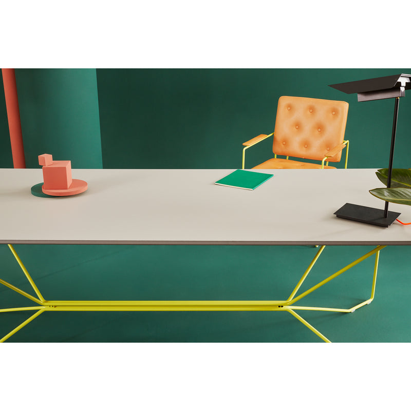A modern office setting showcases the Victor Foxtrot SAME SAME Linoleum Table with a sleek white surface and powder-coated yellow legs. An orange chair, chic black desk lamp, green book, and abstract sculpture complete the look against vibrant green walls.