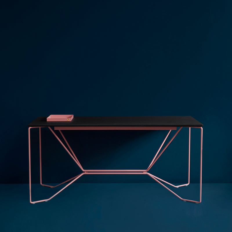 The SAME SAME Linoleum Table by Victor Foxtrot showcases a black tabletop with geometric pink powder-coated steel legs, complemented by a dark blue wall and floor. A closed pink book is placed on the desk.