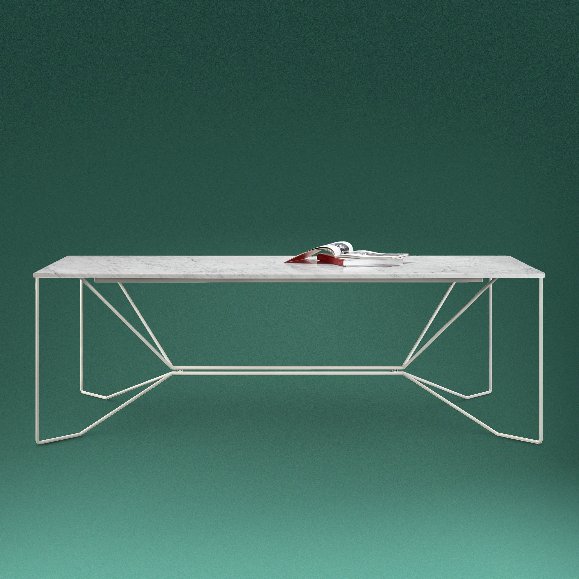 The Victor Foxtrot SAME SAME Marble Table, featuring a sleek white powder-coated steel frame and Carrara Venatino marble top, is set against a rich green backdrop. A red-covered book adds a vibrant color contrast to the modern design.