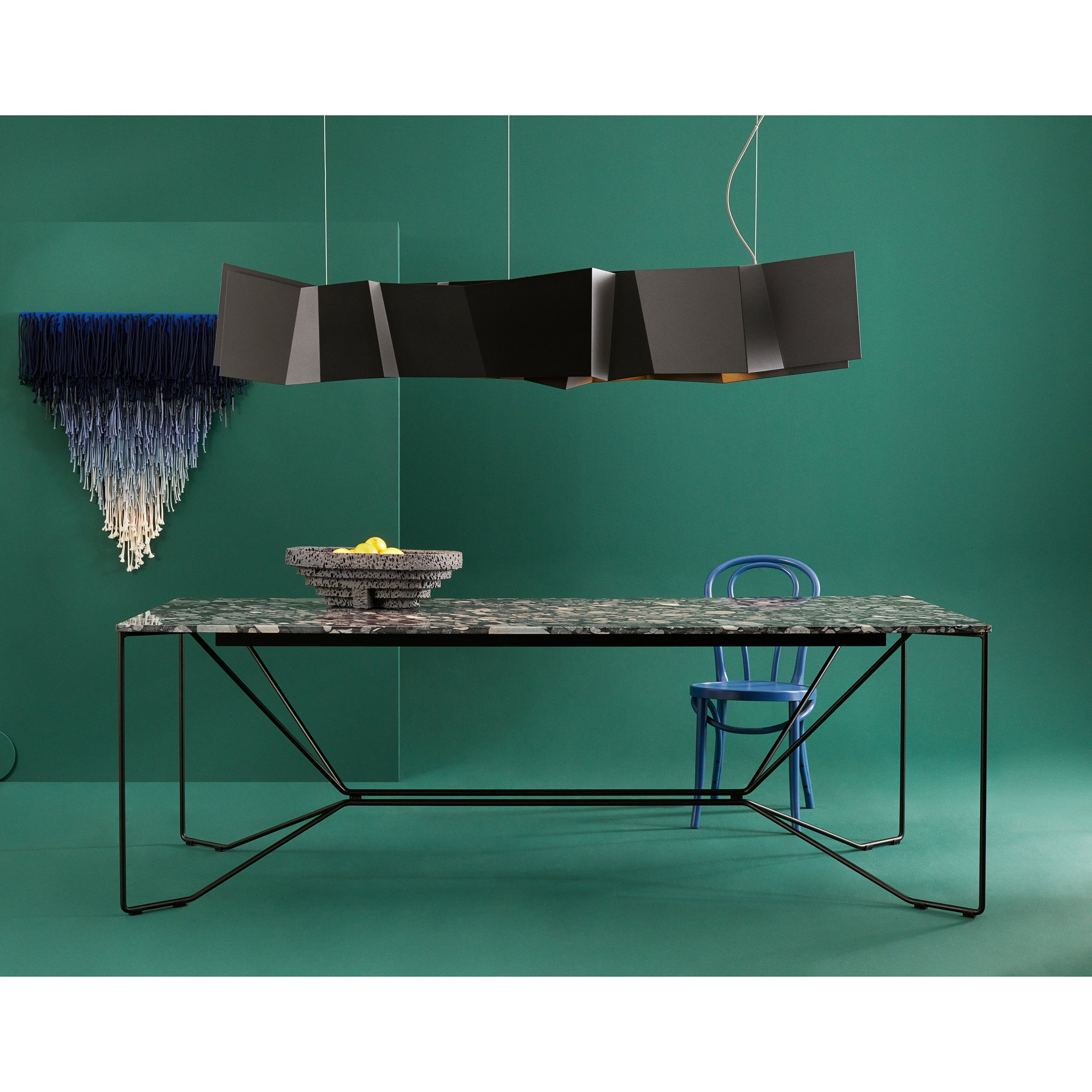 A modern dining setup features the Victor Foxtrot SAME SAME Nero Marinace Table with a geometric metal base in powder-coated steel, set against green walls. A blue chair is under the table, and an angular black pendant light complements the blue and white wall hanging.