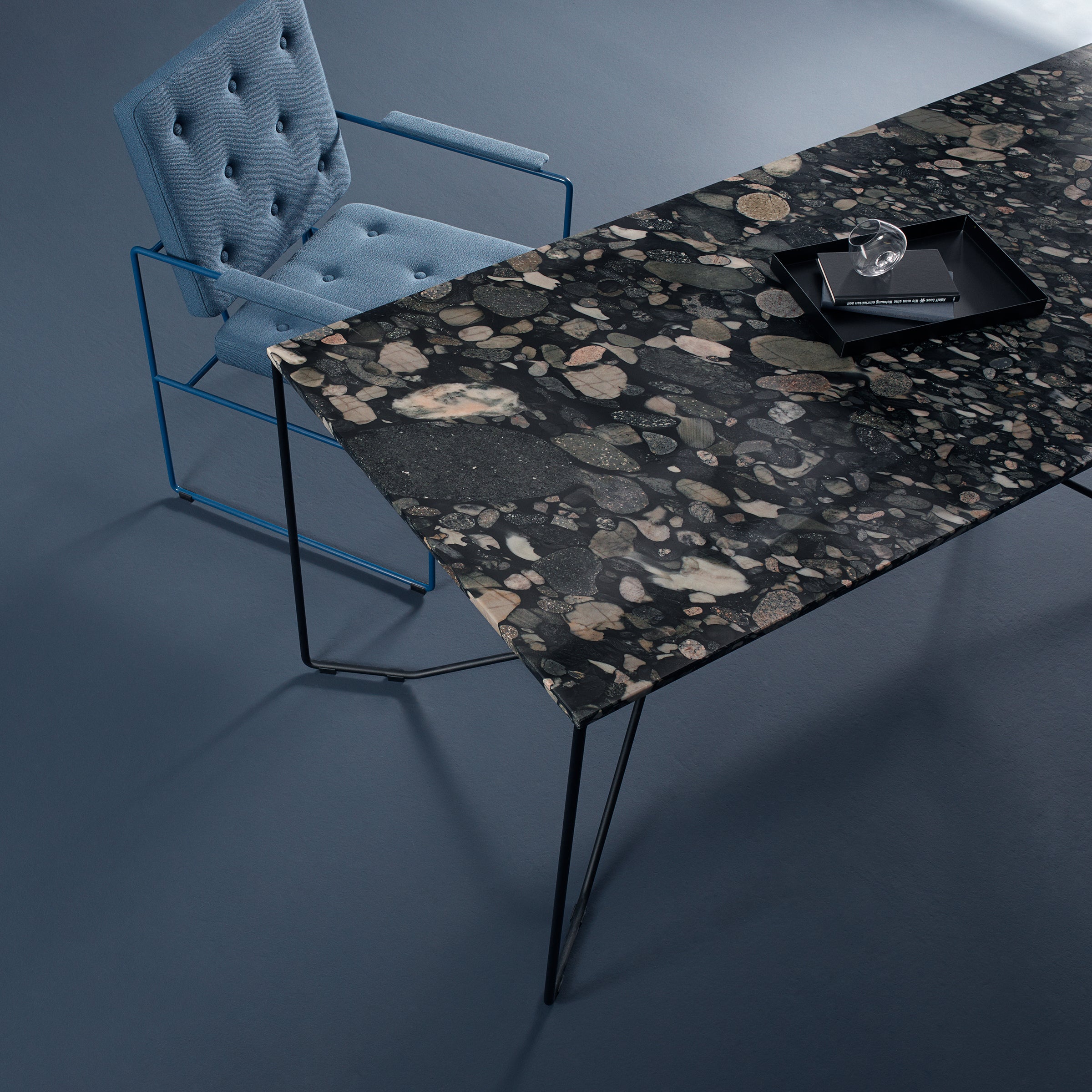 An overhead view of a chic office setup showcases a modern blue chair with a powder-coated steel frame beside the SAME SAME Nero Marinace Table by Victor Foxtrot, featuring a dark, polished stone surface with embedded stones and an elegant black tray on top.