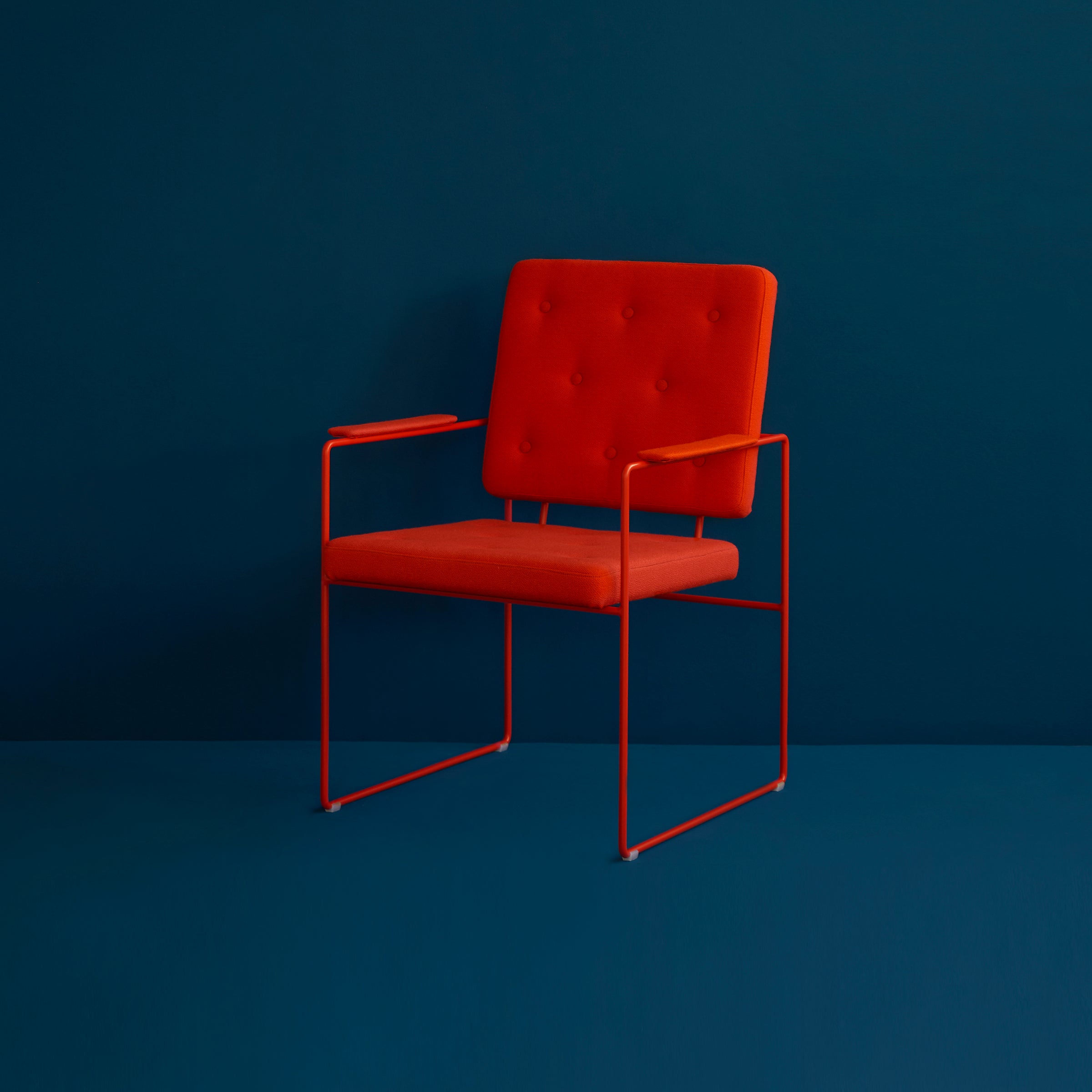 The SWELL TIME Dining Chair by Victor Foxtrot is a minimalist red chair with button-tufted cushioning on the backrest and seat, elegantly supported by slender steel frame armrests and legs, set against a dark blue background.