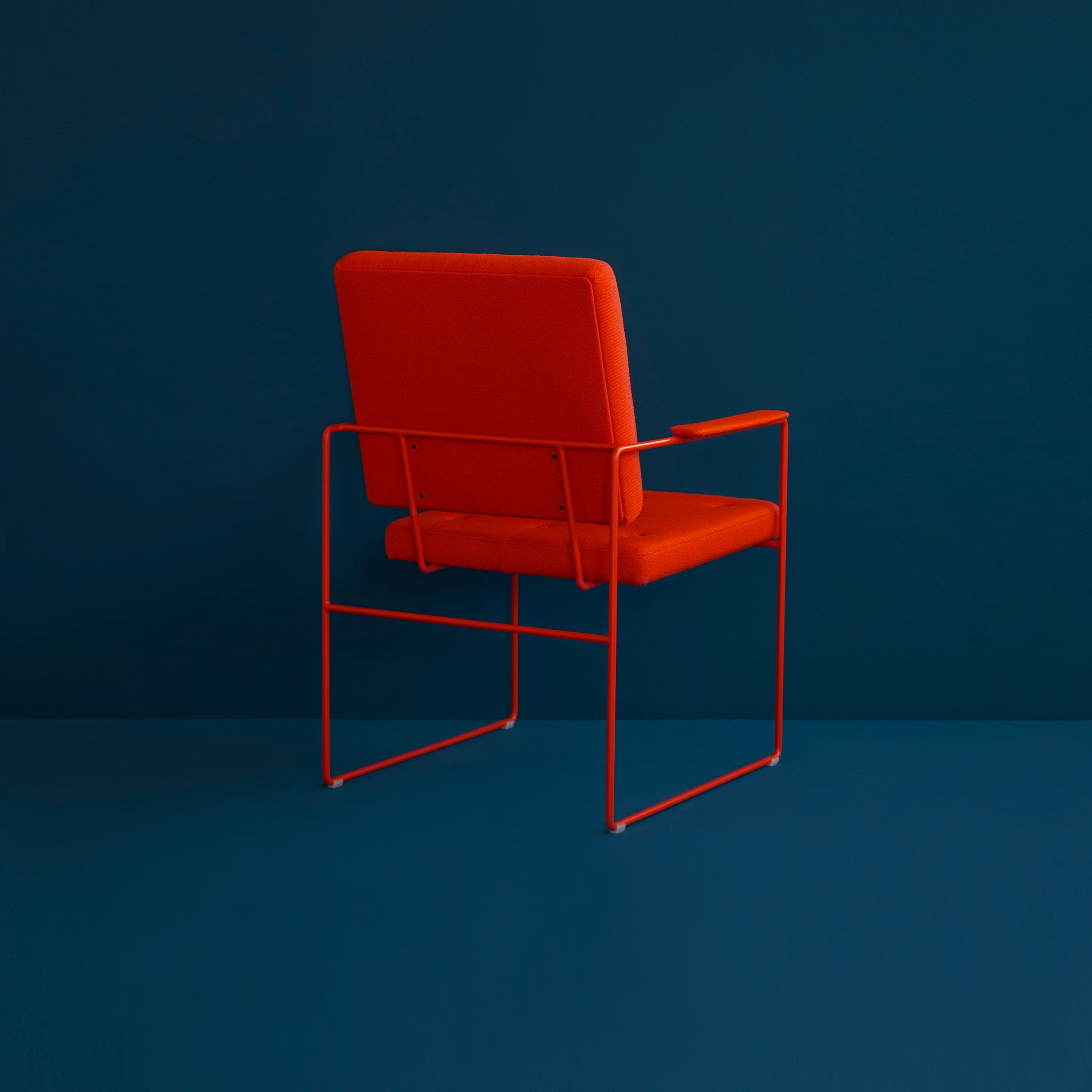 The SWELL TIME Dining Chair by Victor Foxtrot features a vibrant orange cushion and slender steel frame. Set against a dark teal background, the chairs minimalist design includes button stitching that adds subtle texture, with the chair facing away from the viewer.