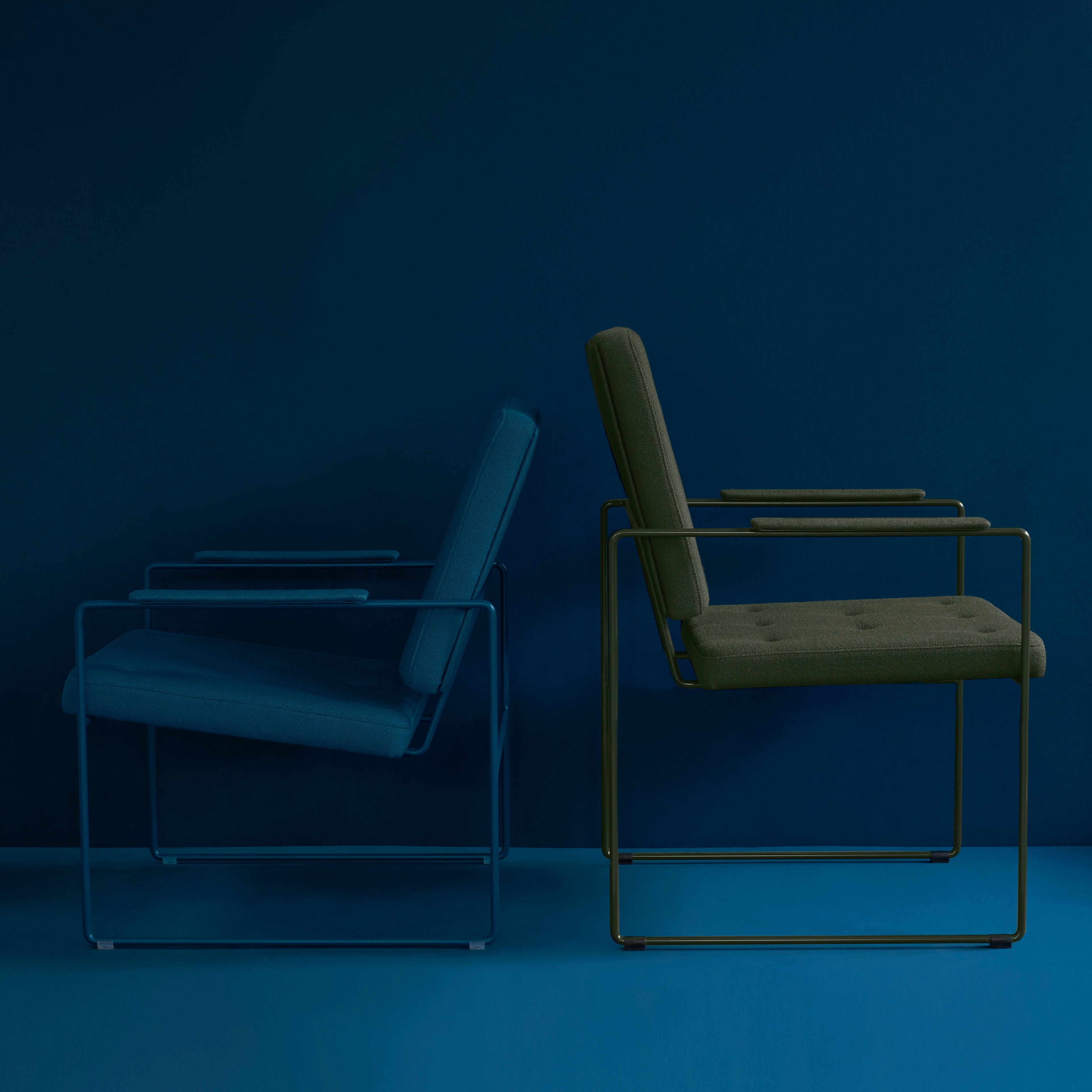 Two SWELL TIME Dining Chairs by Victor Foxtrot stand against a blue backdrop, each featuring a sleek metal frame. One has a blue cushion, the other dark green, reflecting British upholsterys classic boxy design.