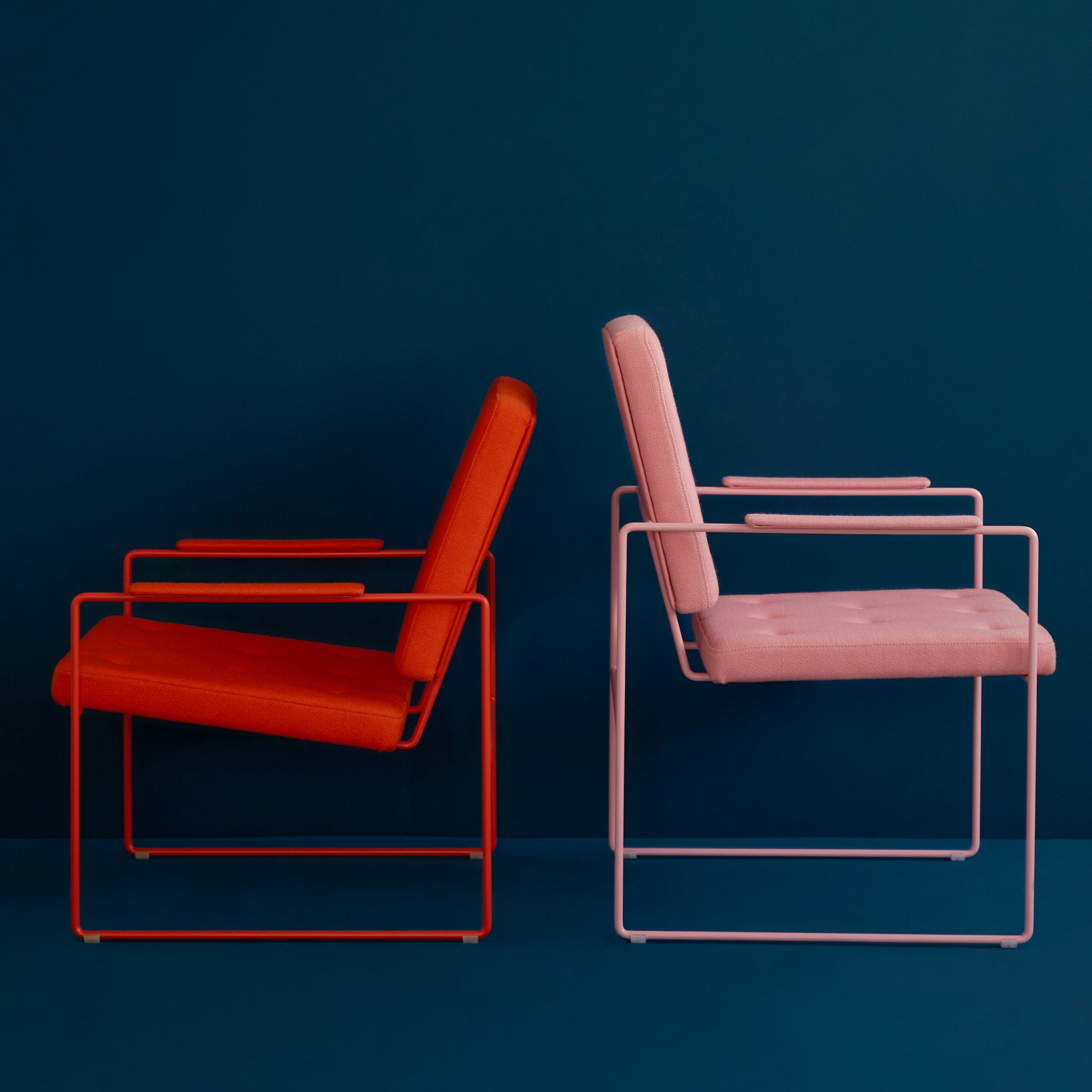 Two SWELL TIME Dining Chairs by Victor Foxtrot are positioned back to back against a dark blue background. One is red with a matching steel frame, and the other is light pink with a matching metal frame, both boasting sleek geometric designs that showcase their contemporary flair.