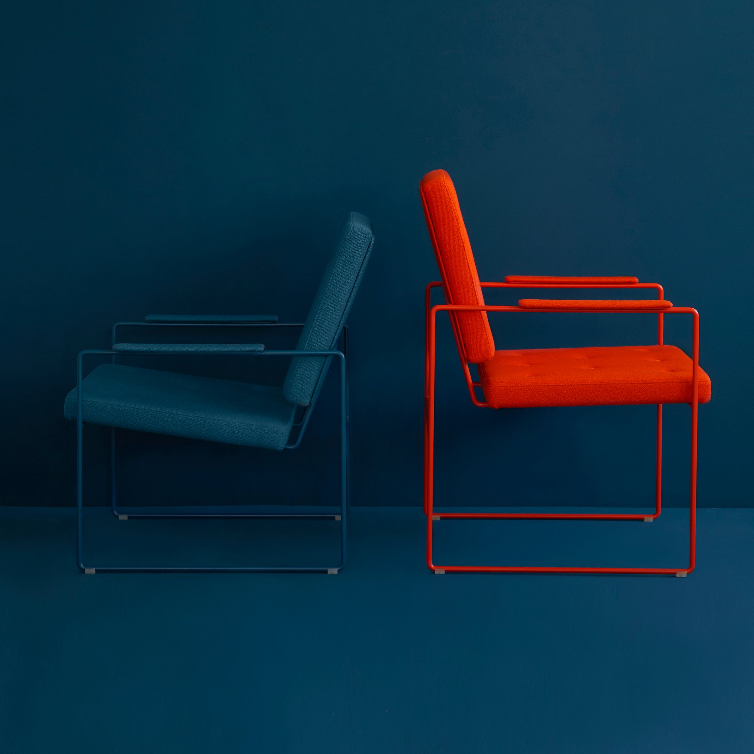 Two SWELL TIME Dining Chairs by Victor Foxtrot with modern steel frames and cushioned seats sit side-by-side. The dark blue chair has subtle button stitching, while the vibrant red one has a sleek finish, both against a matching colored wall and floor for a harmonious look.