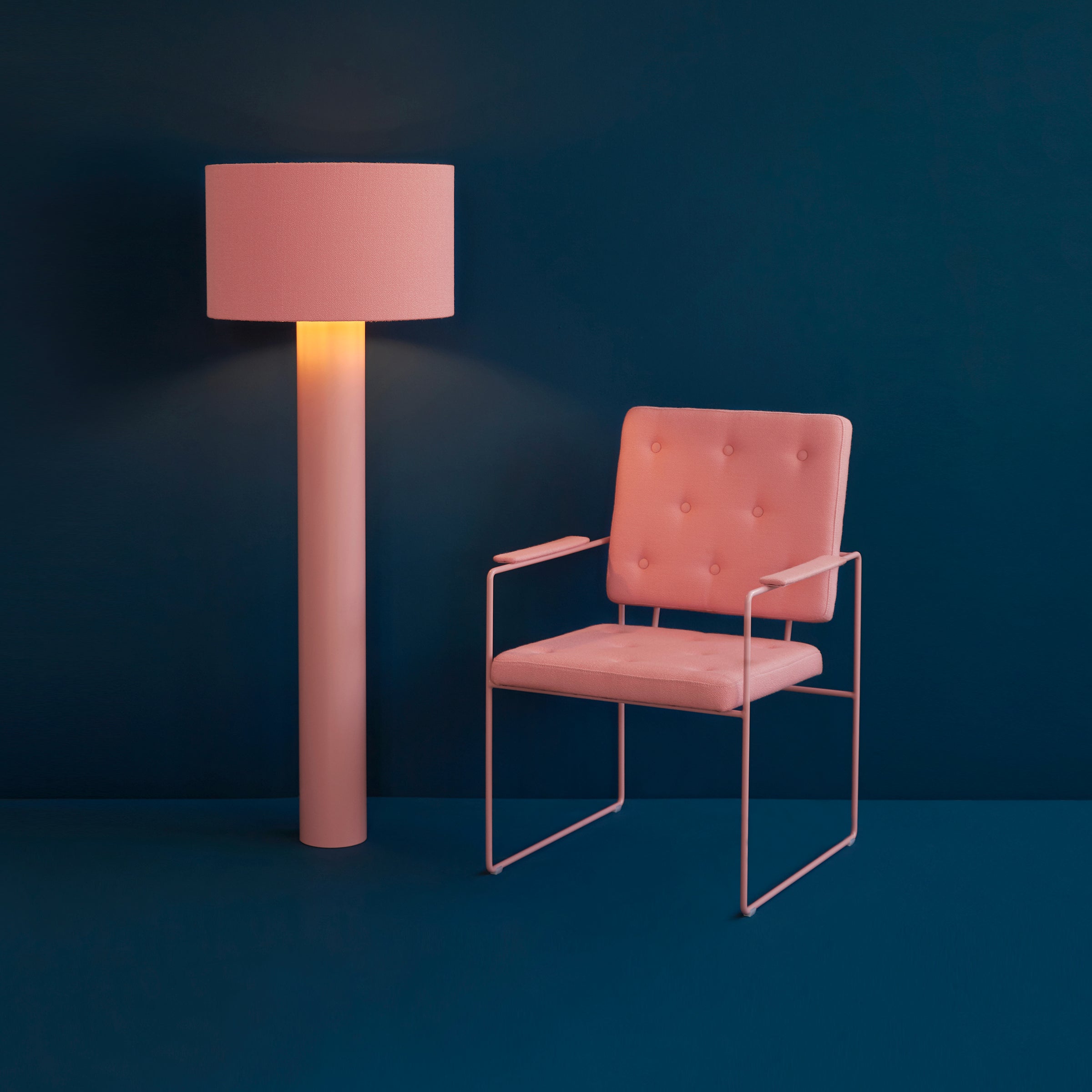 A minimalist scene highlights a SWELL TIME Dining Chair by Victor Foxtrot, featuring pink cushions with button stitching and a sleek steel frame. Next to it is a pink floor lamp with a cylindrical shade, all set against a dark blue background to emphasize the color contrast.