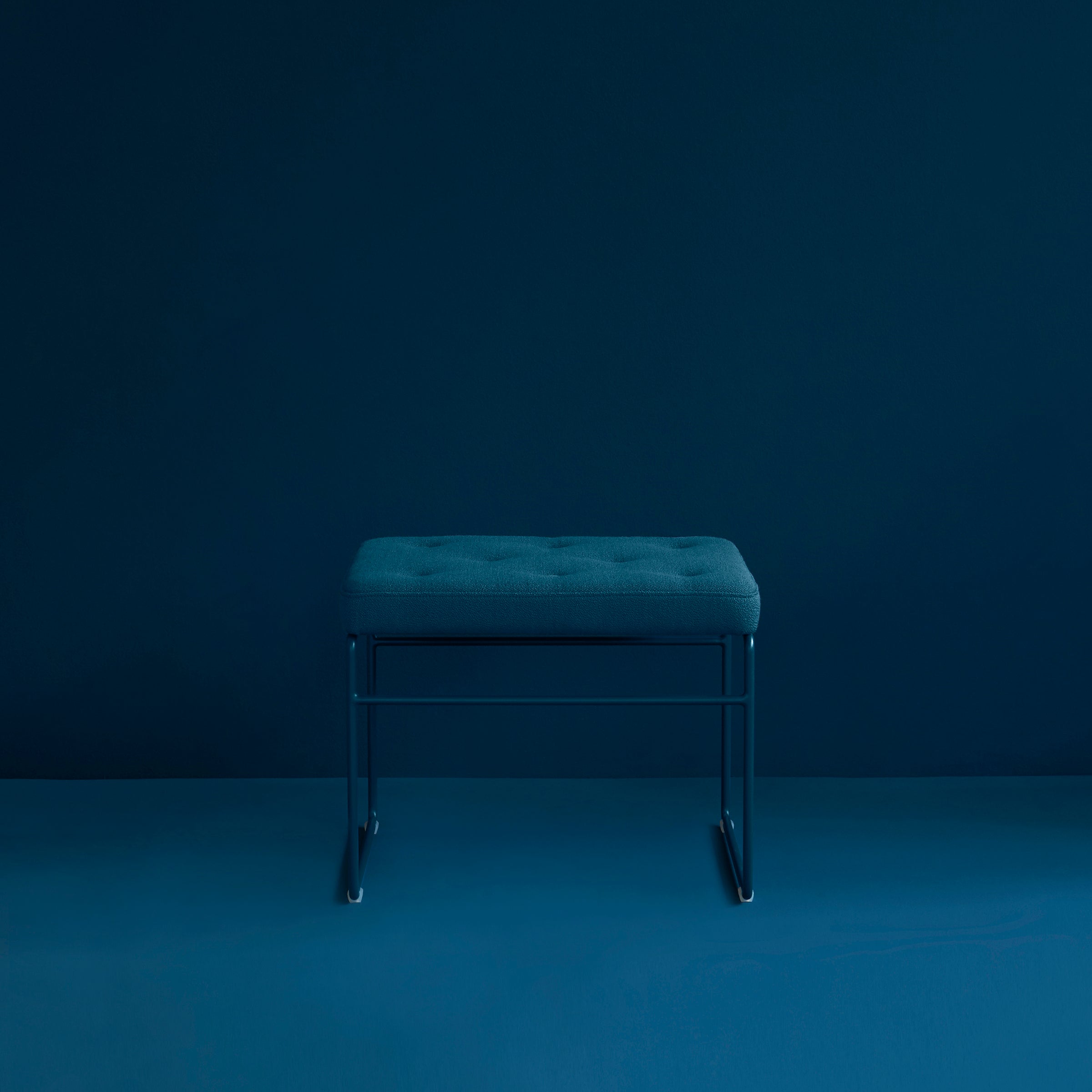 The Victor Foxtrot SWELL TIME Ottoman, with its teal fabric and button stitching on a steel frame, is set against a dark teal backdrop. Its minimalist design and uniform color scheme create a calm, modern atmosphere.