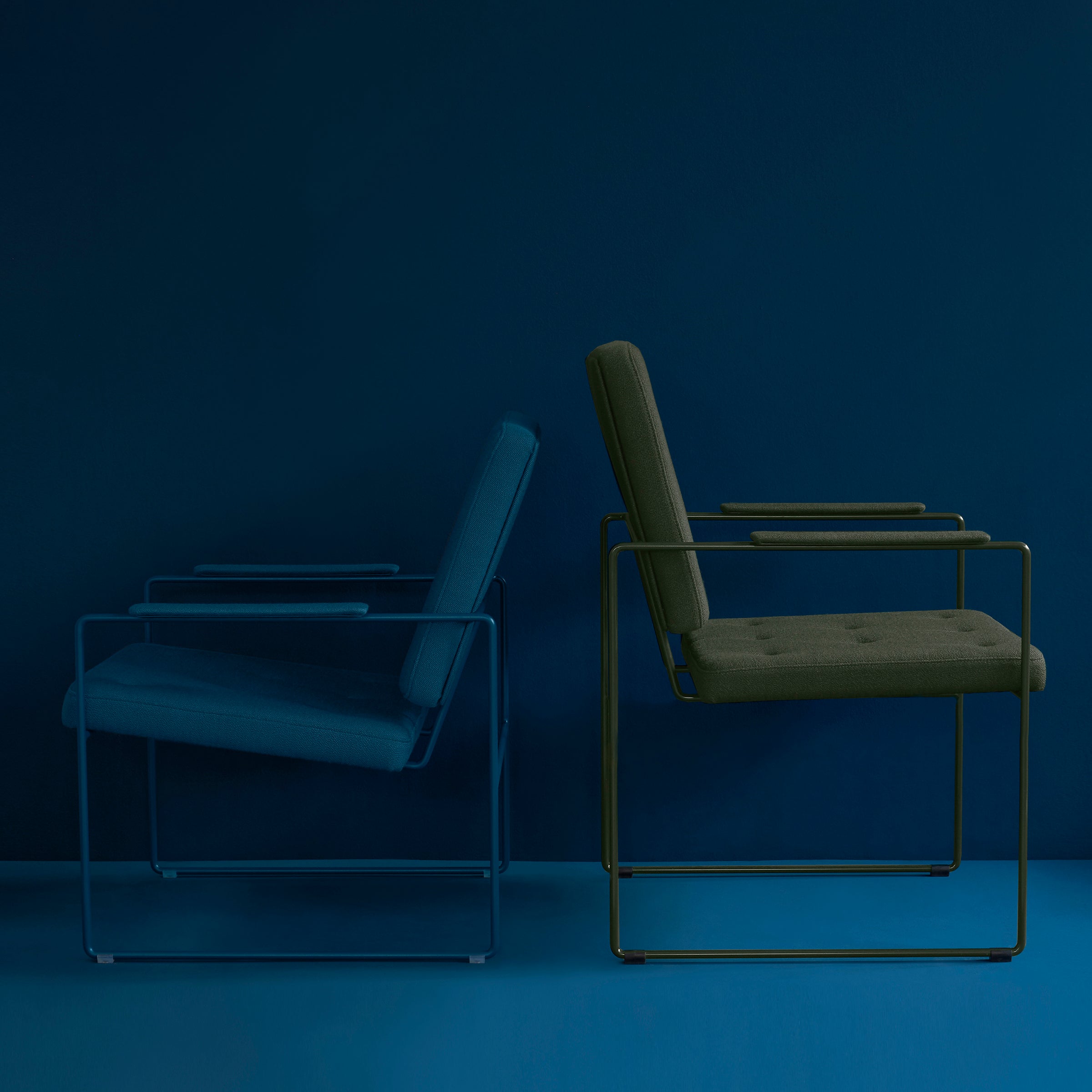 Two SWELL TIME Lounge Chairs by Victor Foxtrot, featuring minimalist designs with sleek steel frames and cushioned seats, one in dark blue and the other in olive green, sit against a deep blue backdrop, exemplifying elegant British upholstery.