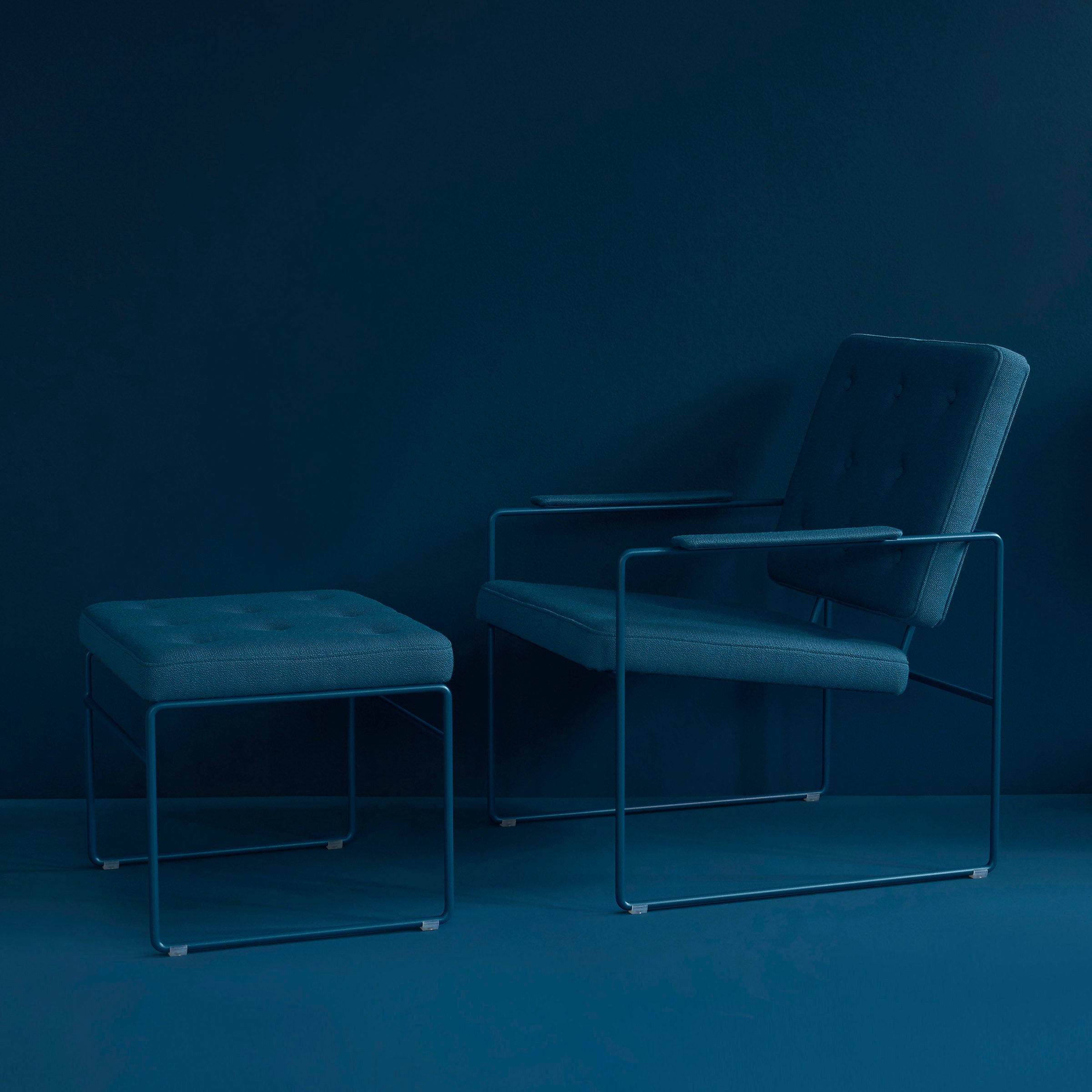 The SWELL TIME Lounge Chair by Victor Foxtrot, with a matching ottoman, features British upholstery and slim metal legs. Seated against a teal wall and floor, this designer piece exemplifies minimalist elegance with simple cushions and monochromatic appeal.