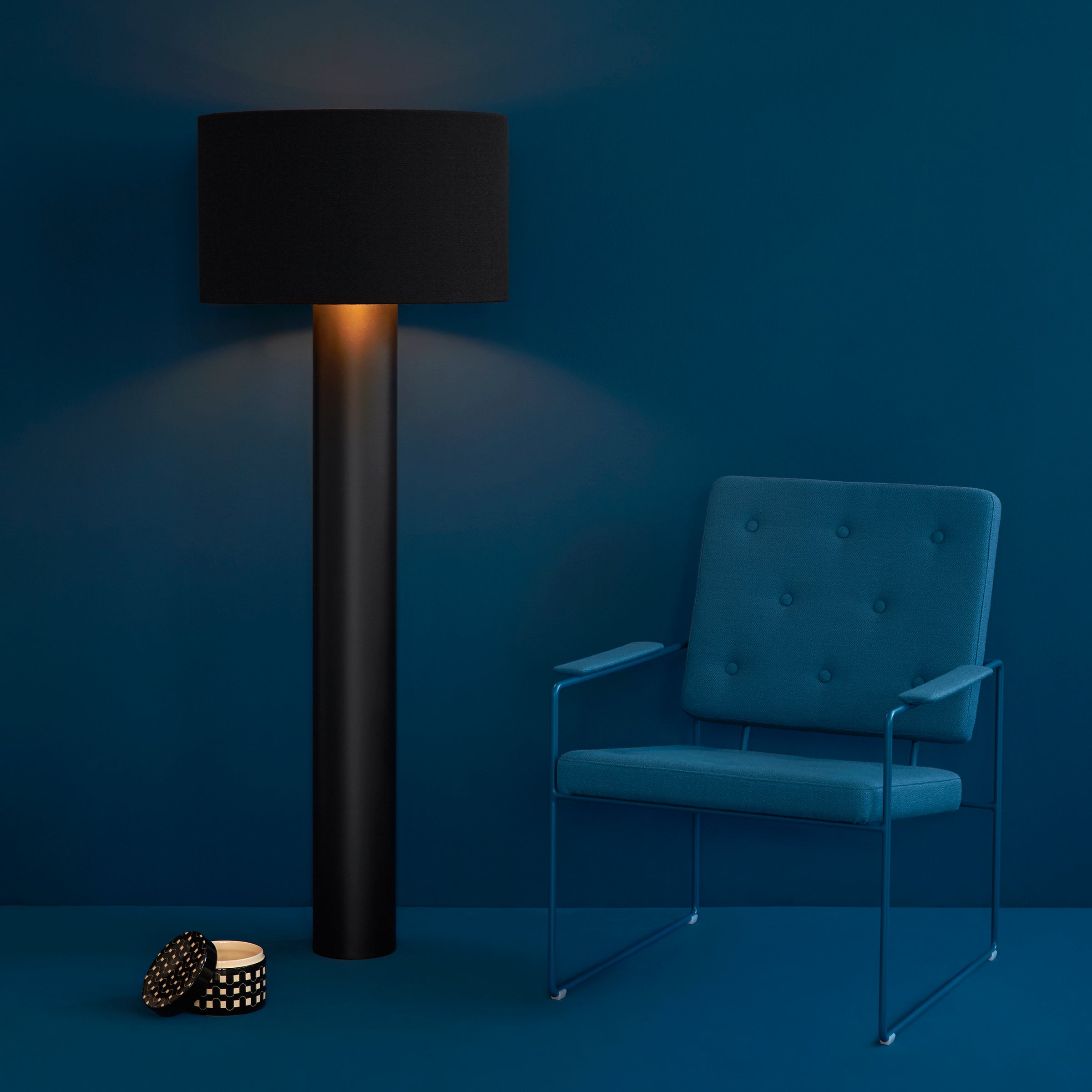 A modern minimalist room features a navy blue wall, a tall black floor lamp with soft light, and the Victor Foxtrot SWELL TIME Lounge Chair with elegant button stitching upholstery and a sleek steel frame. A small decorative container rests on the floor.