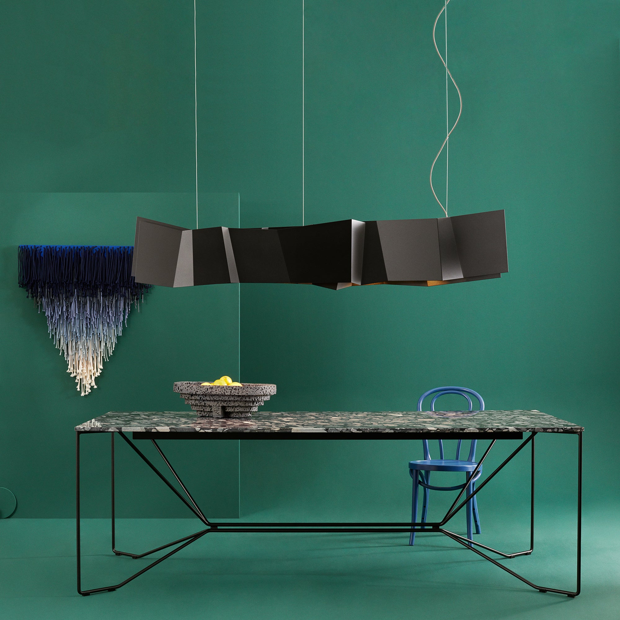 A contemporary dining scene with Victor Foxtrots ZIG ZAG Pendant Lamp over a textured table with a fruit bowl. A blue chair stands nearby. The green backdrop features a stylish blue and white wall decoration.