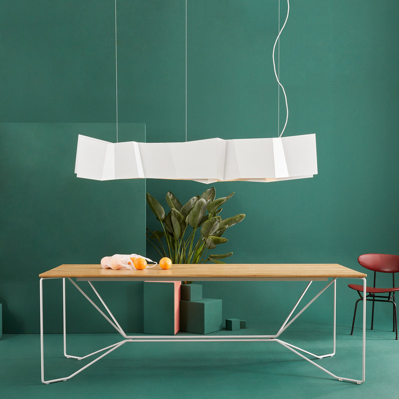 A modern dining room showcases a wooden table with geometric white metal legs and the Victor Foxtrot ZIG ZAG Pendant Lamp overhead. A large plant on the green wall adds nature, while restaurant-style lighting provides elegance. A red chair is partially visible on the right.