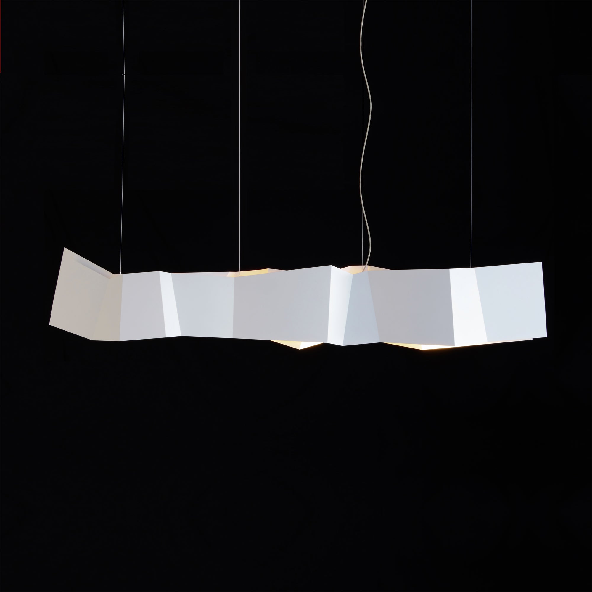 The ZIG ZAG Pendant Lamp by Victor Foxtrot hangs against a black backdrop, showcasing its origami-inspired design with folded geometric shapes. Ideal for restaurants, it suspends by thin wires and emits a soft glow from within the folds.