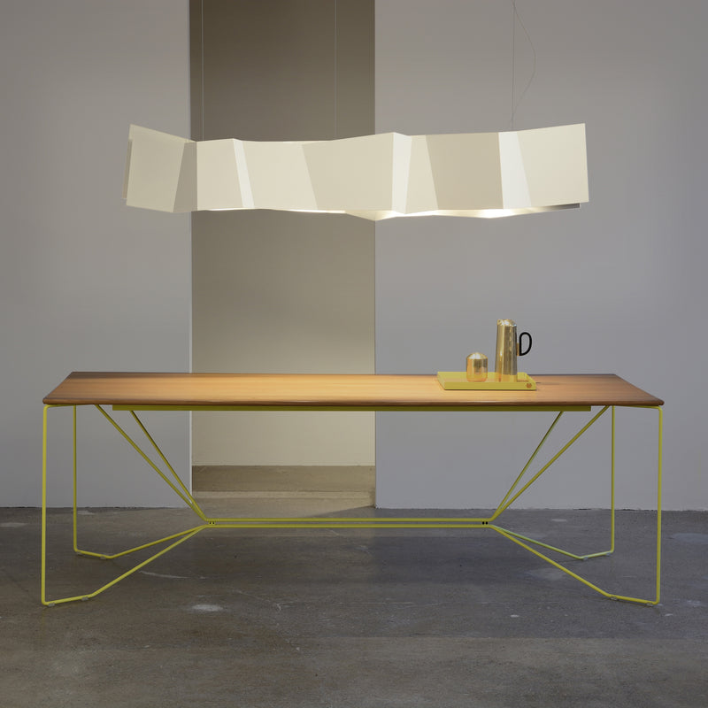 A modern dining table with a slim wooden top and a minimalist yellow metal frame is beautifully lit by the ZIG ZAG Pendant Lamp from Victor Foxtrot. The table features a yellow tray with a metallic pitcher and glass, all set on concrete flooring.