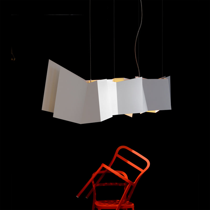 A red chair leans backward at an angle against a dark background. Above it, the ZIG ZAG Pendant Lamp by Victor Foxtrot, featuring contemporary irregular folding shapes, casts a warm glow—ideal for sophisticated restaurant lighting.