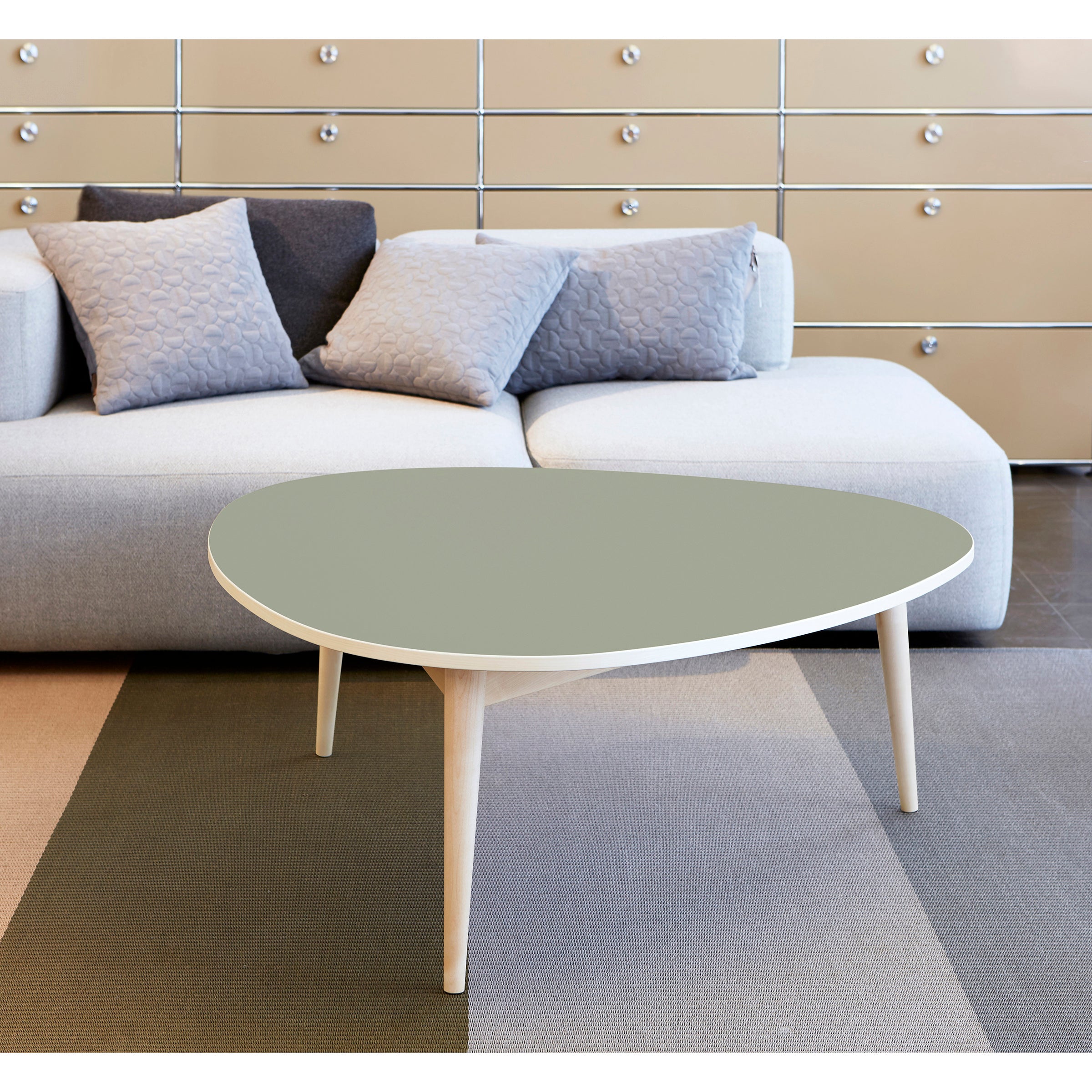 A modern living room features a light-colored sofa adorned with patterned gray and blue cushions. The Drei-Rund Coffee Table by WB Form, with light wooden legs, rests on a textured rug against cream-colored drawers.