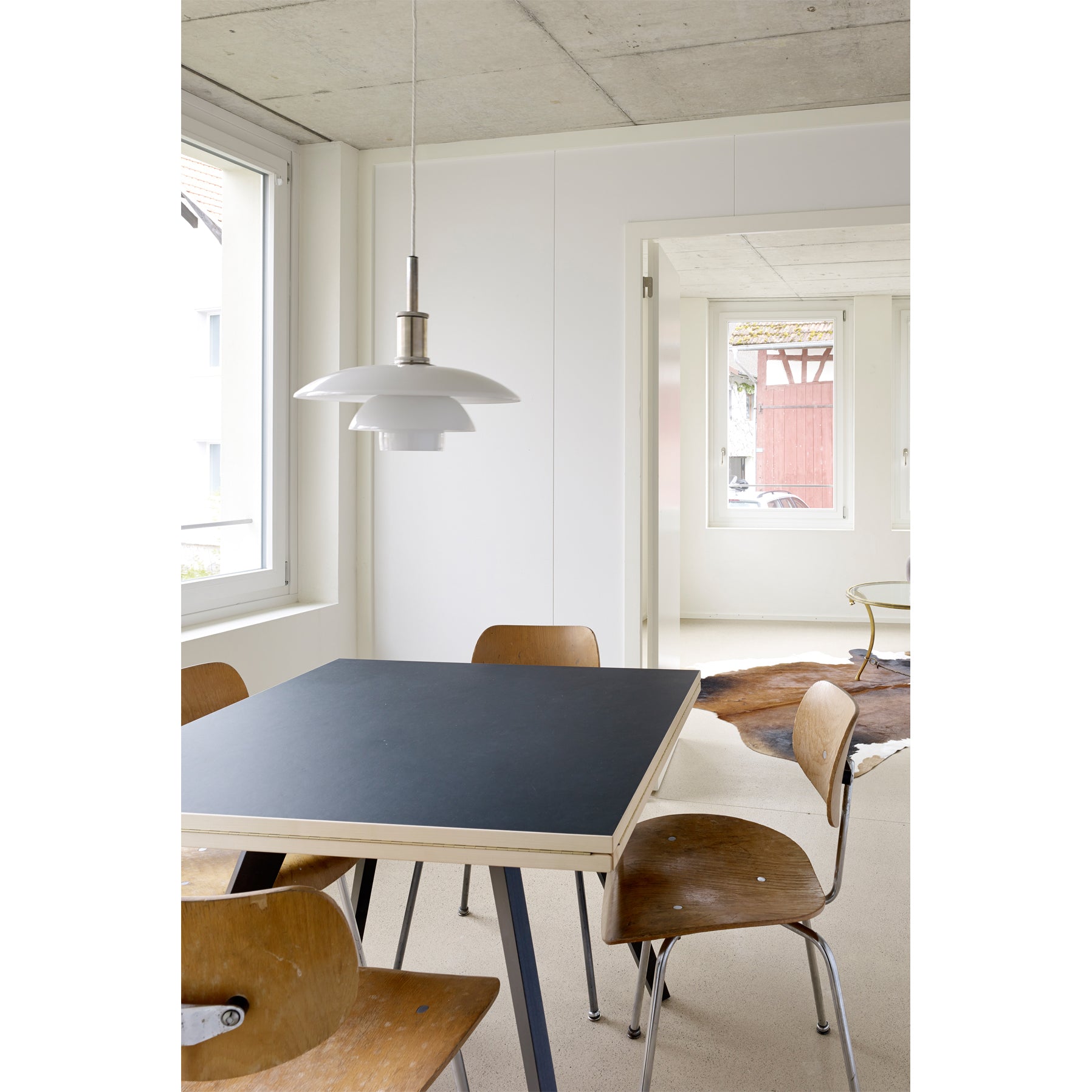 A modern dining room showcases a black WB Form Quadrat-Rund Table with wooden chairs. A pendant light above and large windows bring in natural light, complementing the white walls and concrete ceiling for a chic space inspired by Max Bill design and Wohnbedarf Zurich aesthetics.