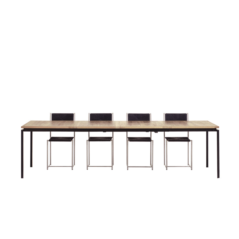 The UPW 1010 Extendable Table by WB Form features a light brown wooden surface and black metal legs. Five black chairs with metal frames are evenly aligned on one side, all set against a plain white background.