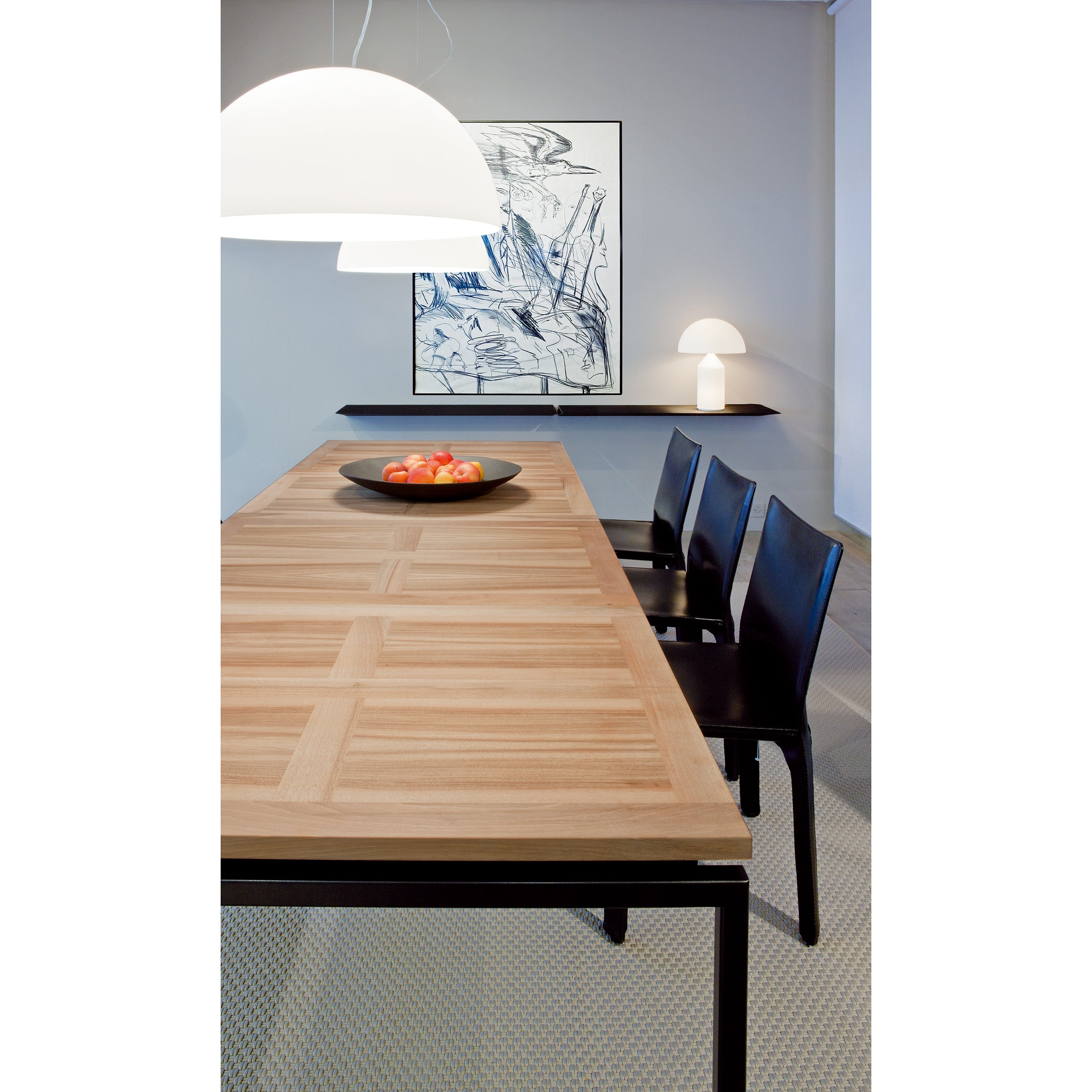The modern dining room features superior workmanship with a long UPW 1010 Extendable Table by WB Form, four black chairs, and a white pendant lamp overhead. A black tray of red apples adds charm, while framed abstract artwork and a table lamp elegantly decorate the back wall.