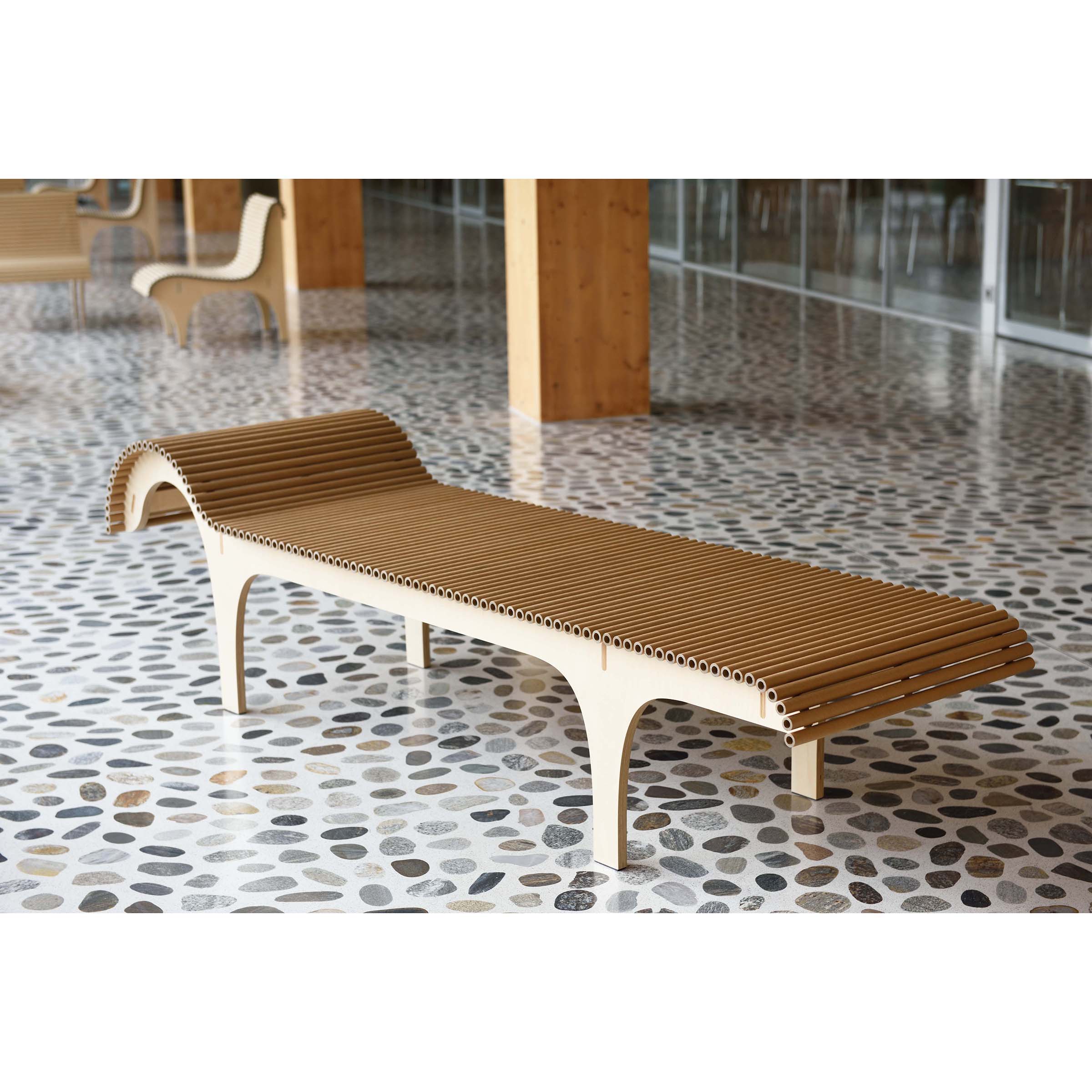 A modern wooden Carta Chaise Longue by WB Form with a slatted design and curved ends rests on a pebble-patterned floor. The space includes large windows, wooden columns, and eco-friendly furniture inspired by Shigeru Bans designs, with another chair and table in the background.