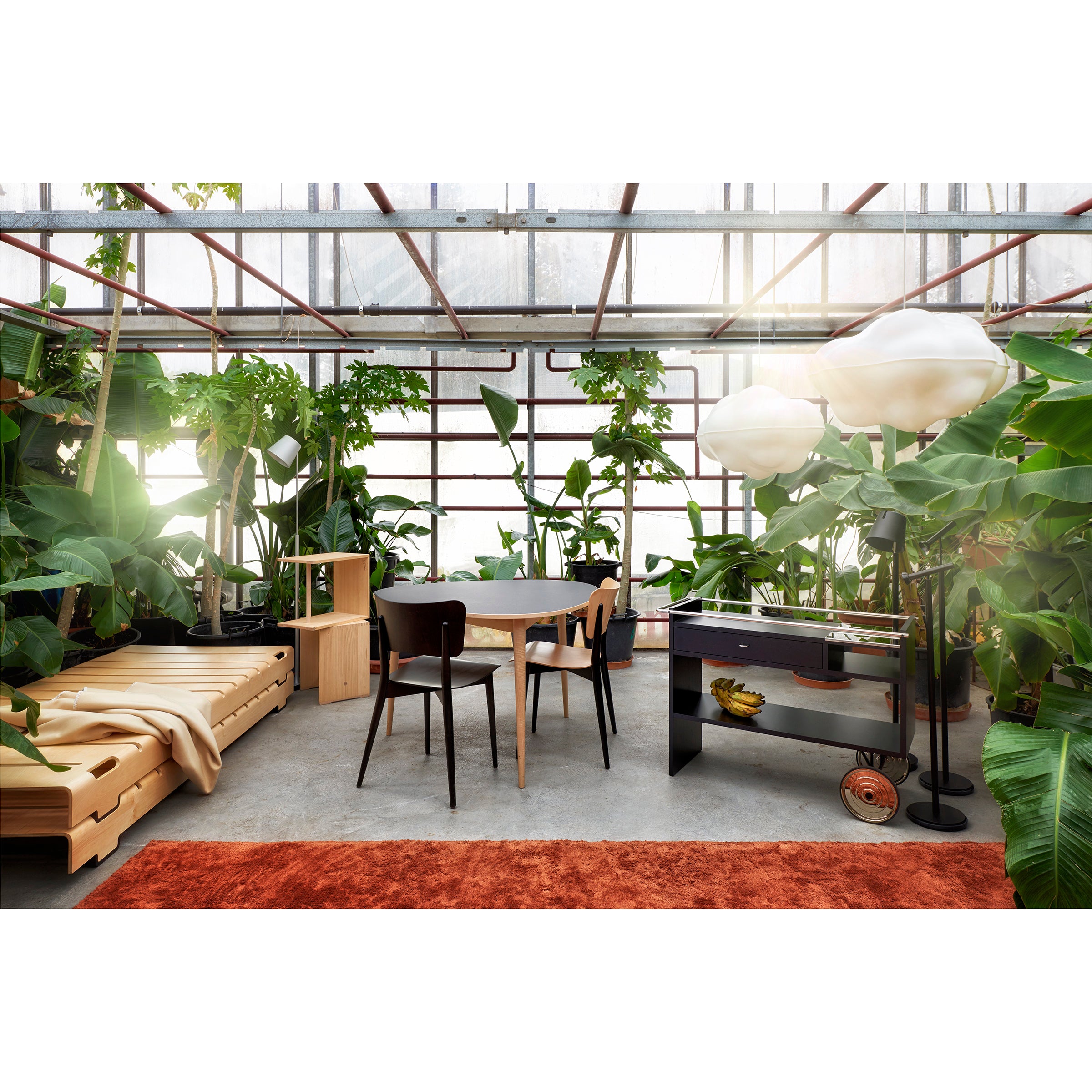 A modern sunlit room features lush green plants, a round wooden table with chairs, and a black sideboard on wheels. A light wood daybed sits beneath Cloud Lamps by WB Form, accompanied by an orange rug. The space is enclosed by a glass and metal framework.
