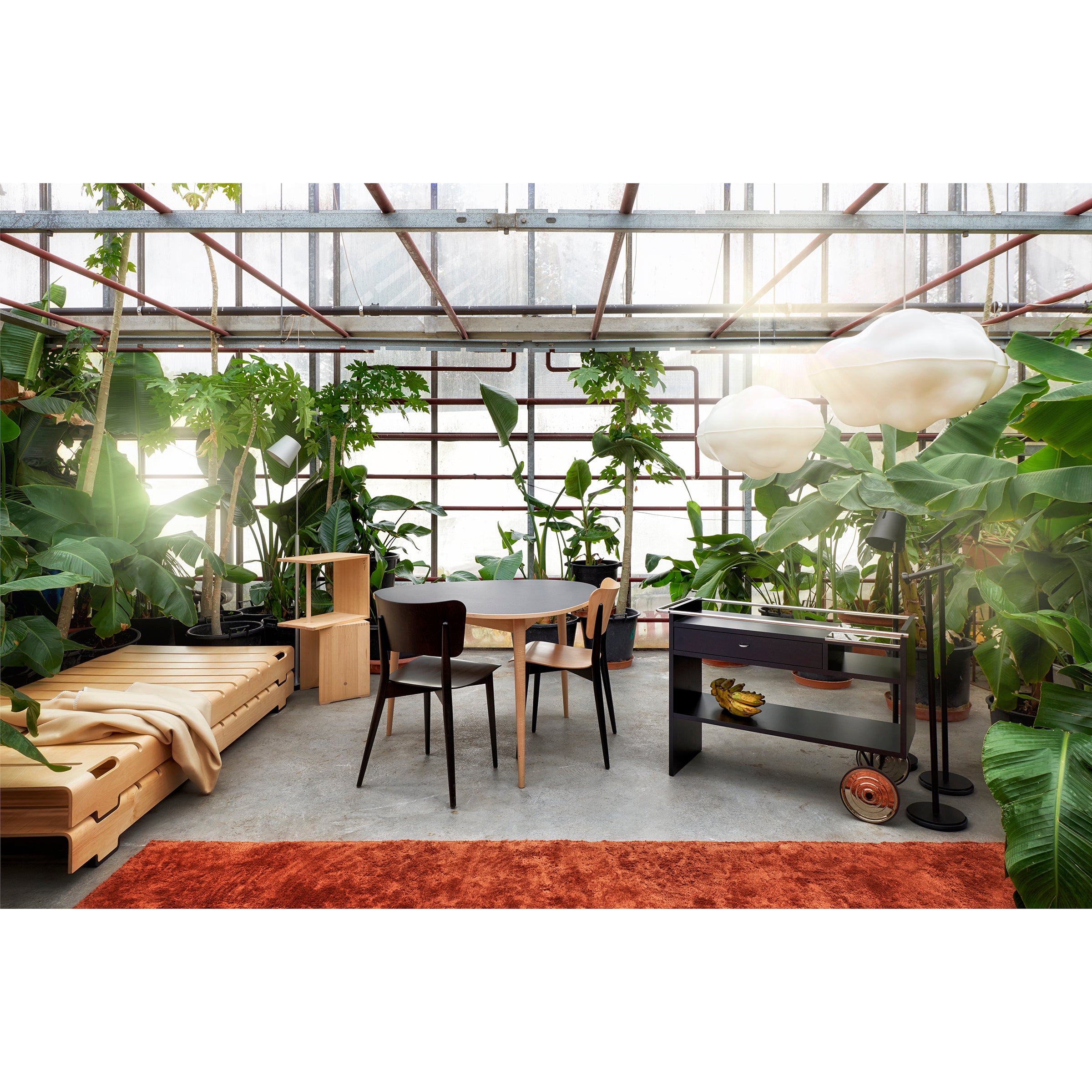 A modern, sunlit room with lush green plants features a WB Form Kreuzzargenstuhl chair. It includes a wooden daybed, black chairs around a round dining table, and a cart with fruits. Cloud-like pendant lights hang above, with an orange rug adding warmth to the concrete floor.