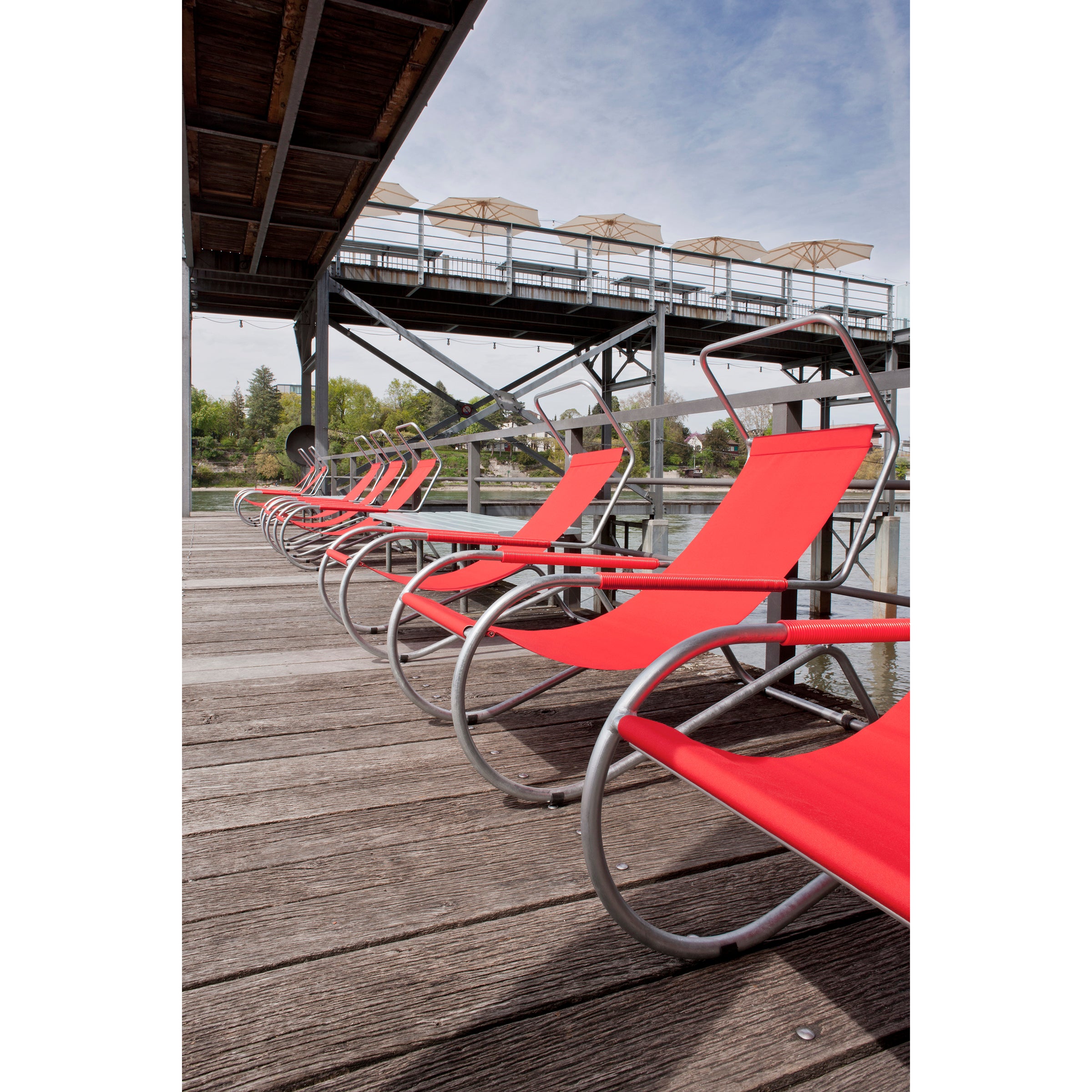 A row of WB Forms Lido Loungers adorns the wooden waterside deck. Overhead, a metal-framed walkway with umbrellas extends to a dock structure. In the background, trees and a building stand under a cloudy sky, completing this tranquil scene.