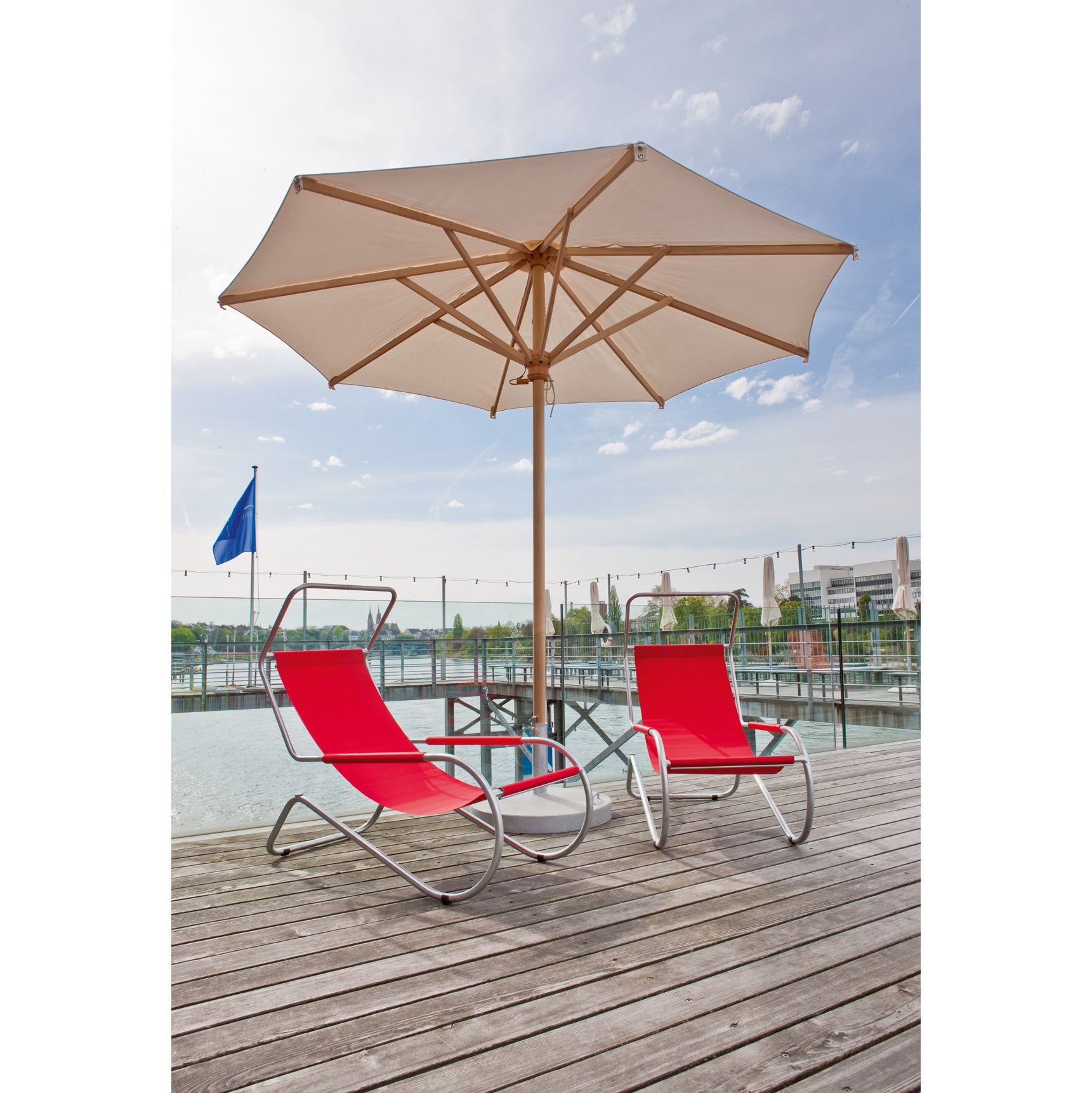 Two red WB Form Lido Loungers with metal frames sit under a large white umbrella on a wooden deck. The deck overlooks water with a visible blue flag, and buildings and trees line the distant shore under partly cloudy skies.