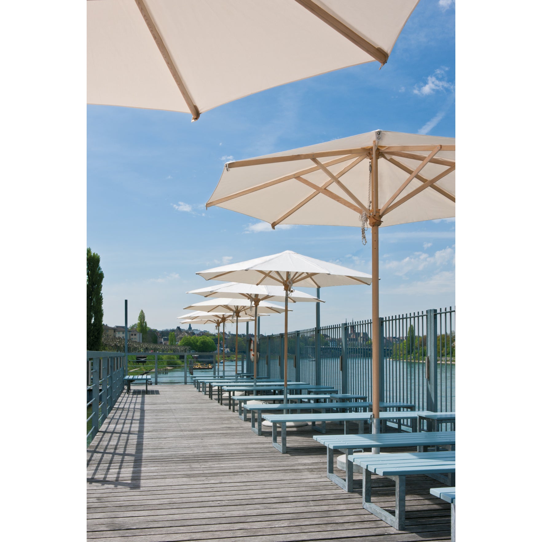 A row of WB Form Ombrelloni parasols shades the blue benches on a wooden boardwalk, enclosed by a fence. The scene is against a clear blue sky with distant buildings, hinting at a riverside or waterfront locale.
