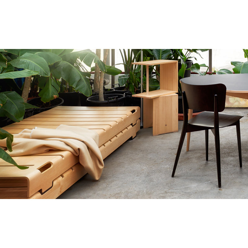 A minimalist room showcases the Turtle Stacking Bed by WB Form, complemented by a small side table and chair. A beige blanket drapes over the bed, while large green plants surround the flexible furniture, creating a refreshing natural atmosphere.