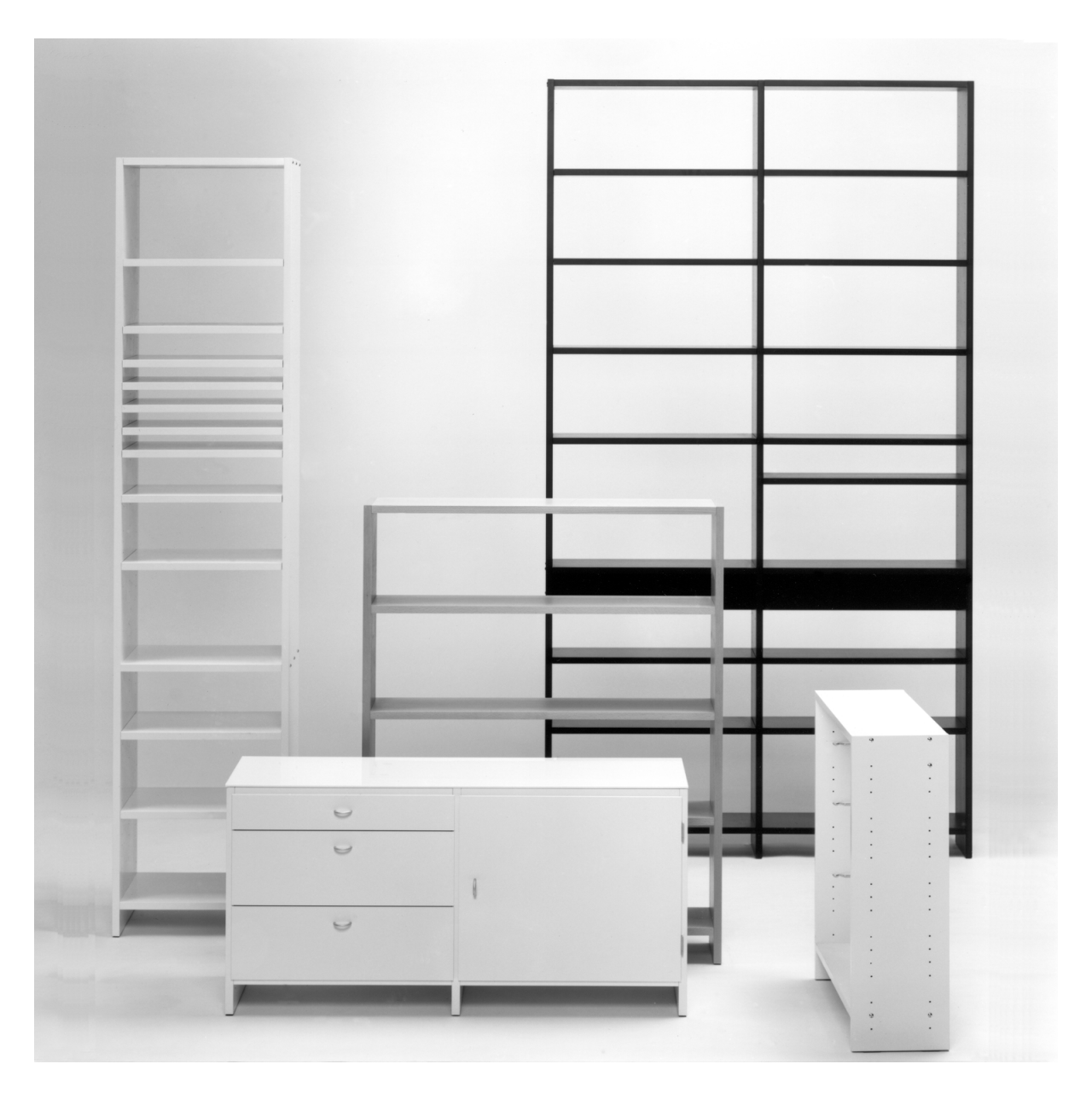 A black and white image displays WB Forms UPW Shelf: a modern system of three tall, narrow bookshelves—one black, two white—alongside a lower white cabinet with drawers, embodying simplicity against a plain white background.